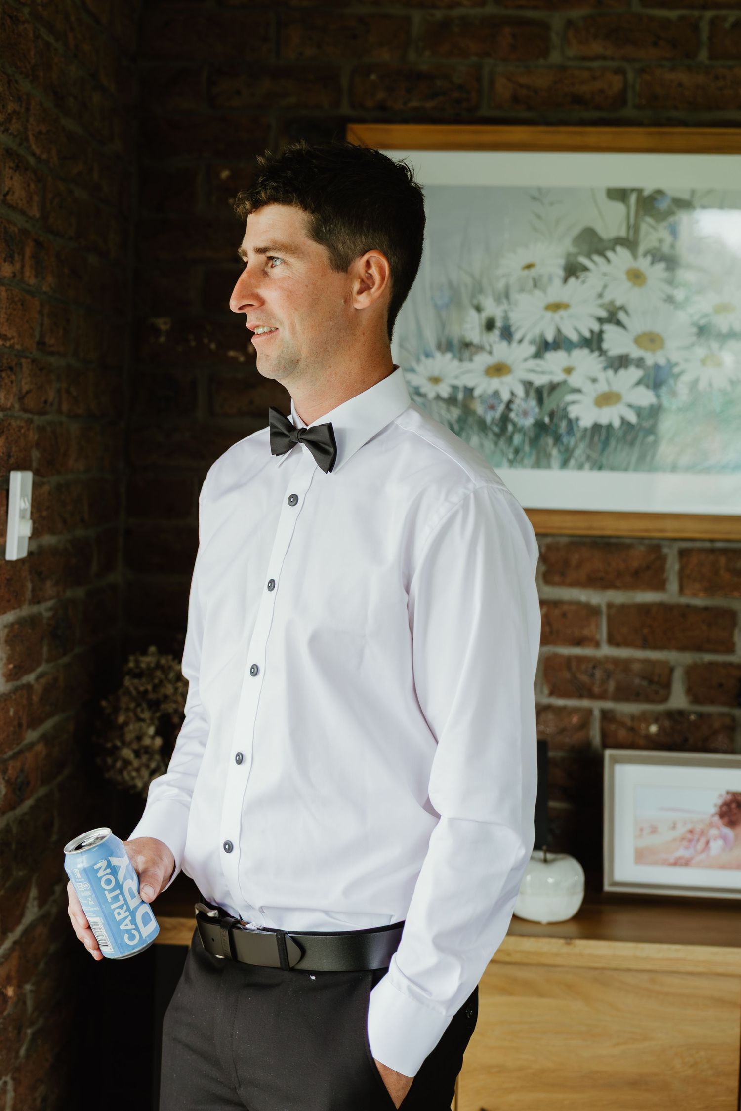 Dapper groom in white shirt and bow tie holds a drink can beside brick wall.