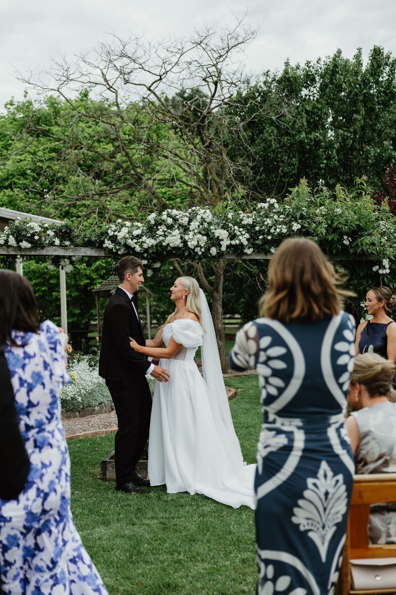 Bride and groom exchange vows in lush outdoor garden ceremony with white floral arch.