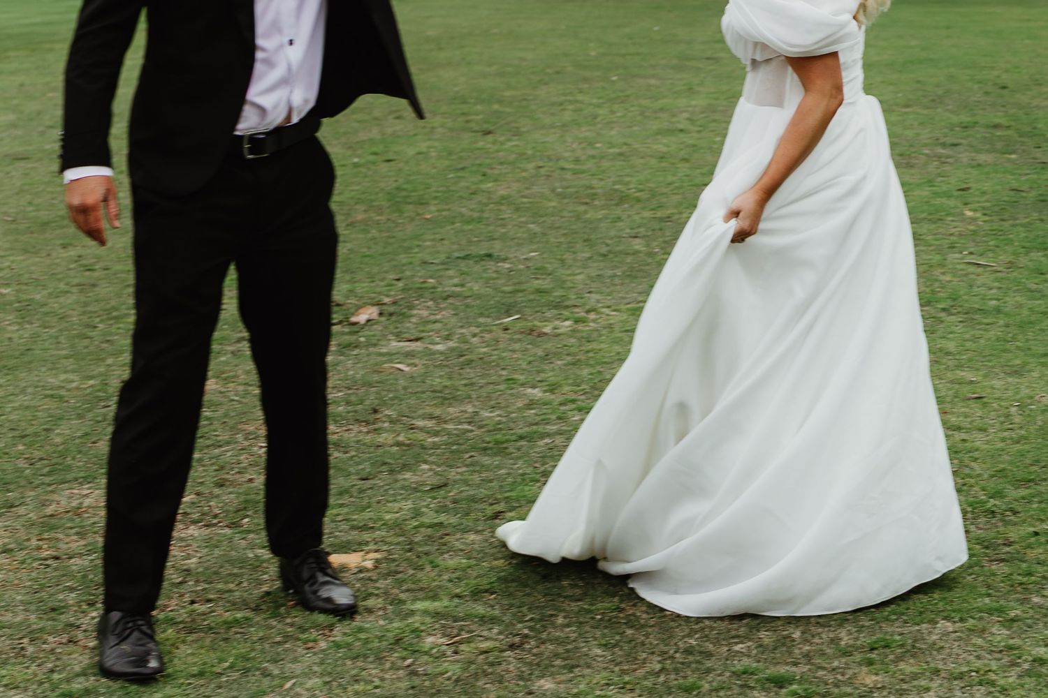 Bride in flowing white gown walks toward groom in black suit on green lawn.