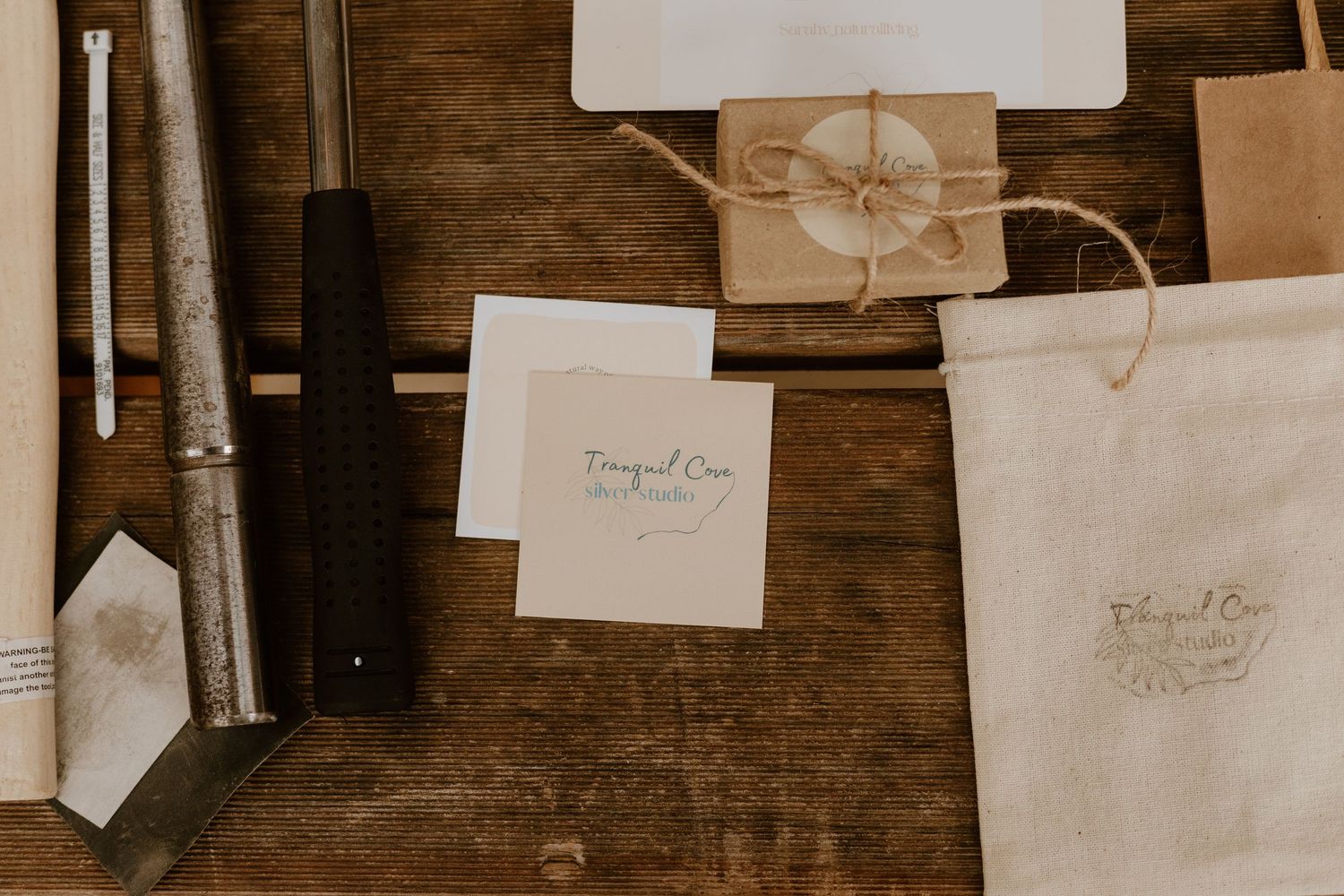 Rustic flat lay of stationery, kraft paper gift box, linen bag, and bookbinding tools on a wooden surface.