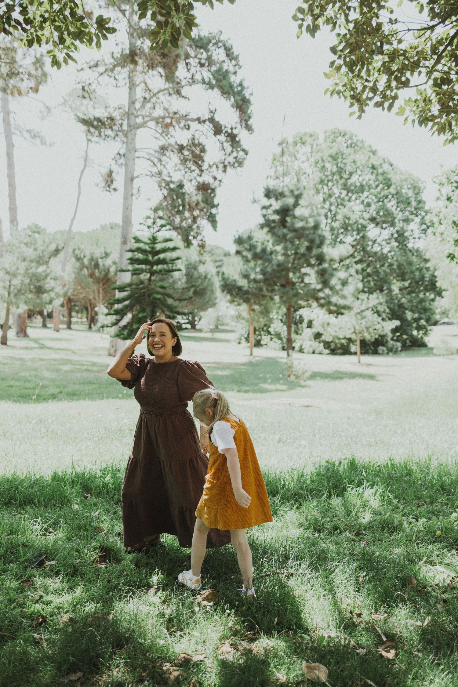 A laughing mother and her young daughter in a yellow dress enjoy a sunny day in a green park.