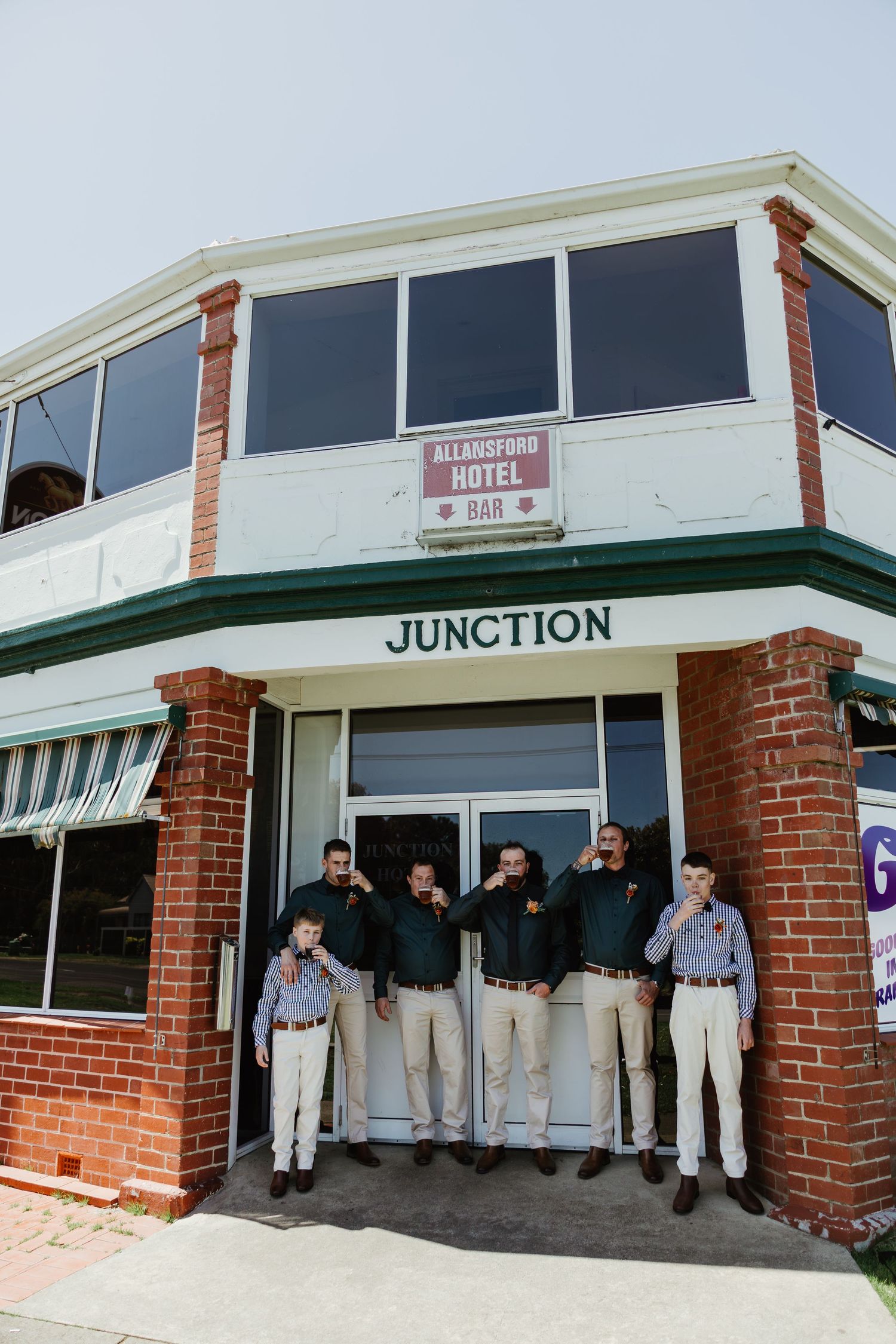 Groomsmen in matching dark shirts and khakis raise drinks outside the Allansford Hotel Junction.