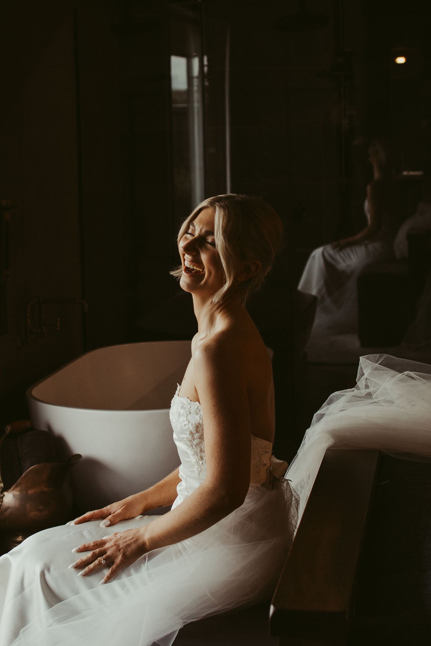 Radiant bride in a strapless lace wedding gown laughing joyfully while seated in a softly lit room.