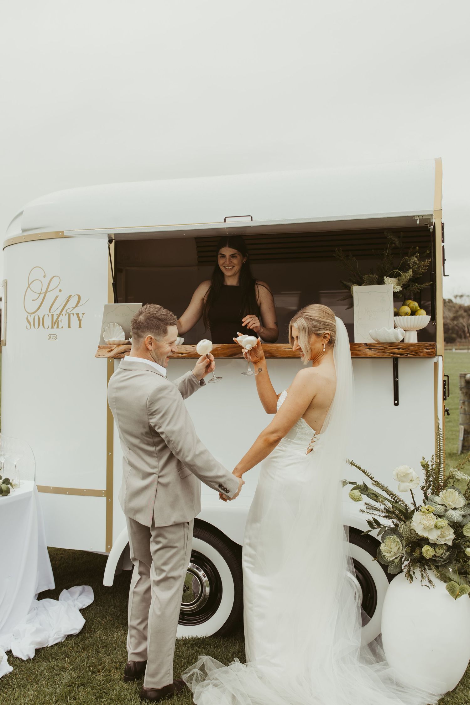 Bride and groom receiving drinks from a white Sip Society bar cart at their outdoor wedding reception.