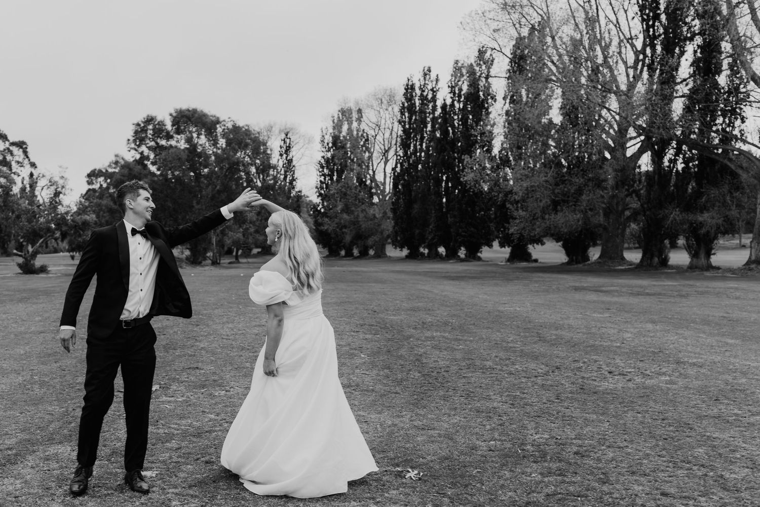 Black and white photo of groom spinning bride in white gown on open grass field with trees.