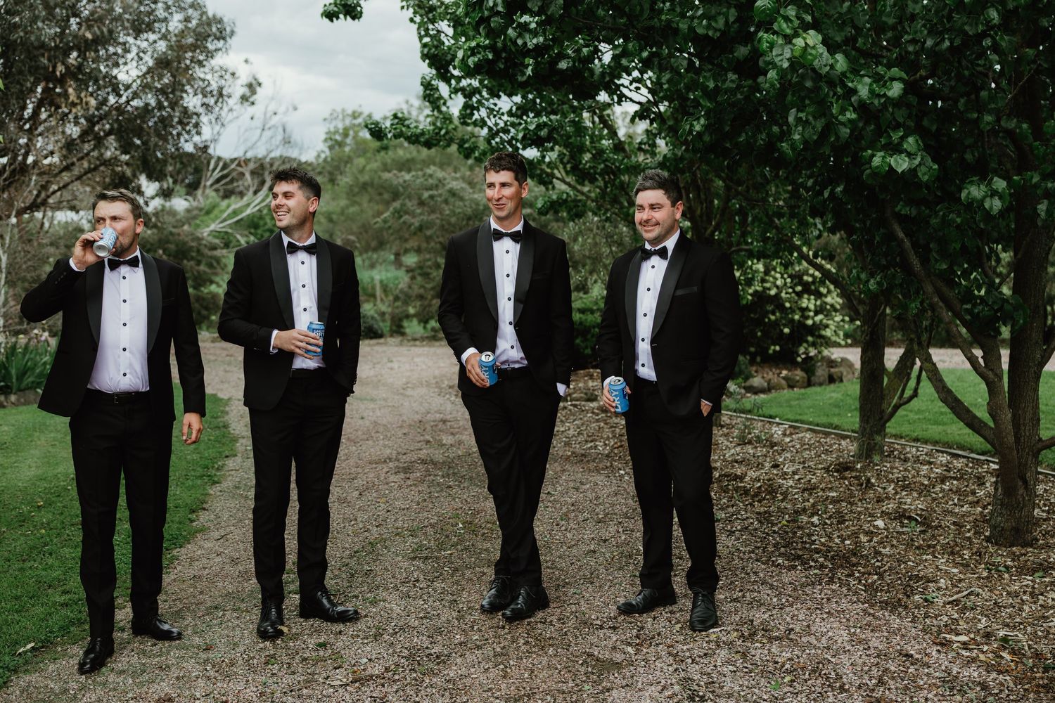 Groom and groomsmen in black tuxedos walking on garden path before wedding.