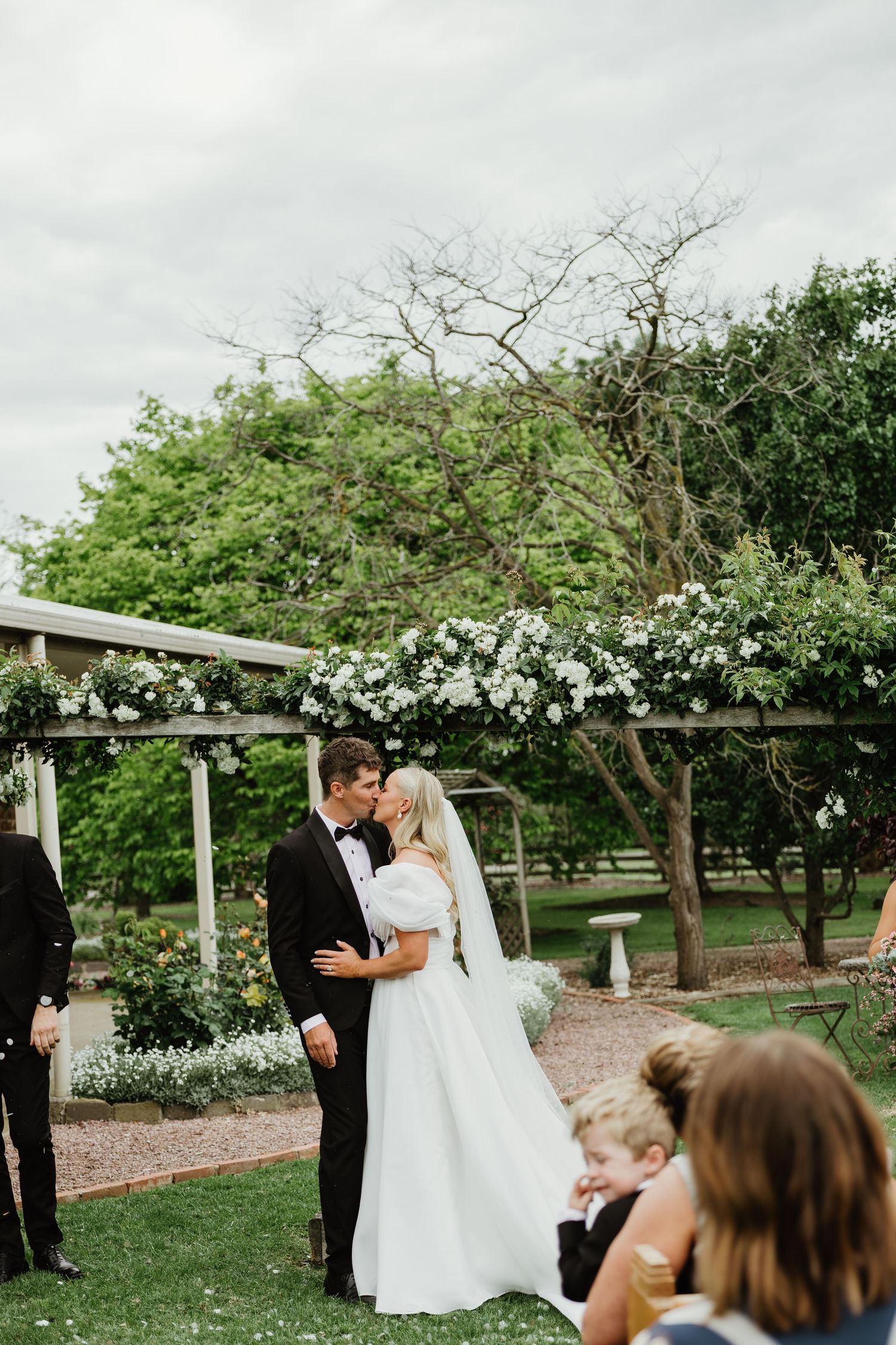 Bride and groom share first kiss under white floral pergola in garden wedding ceremony.