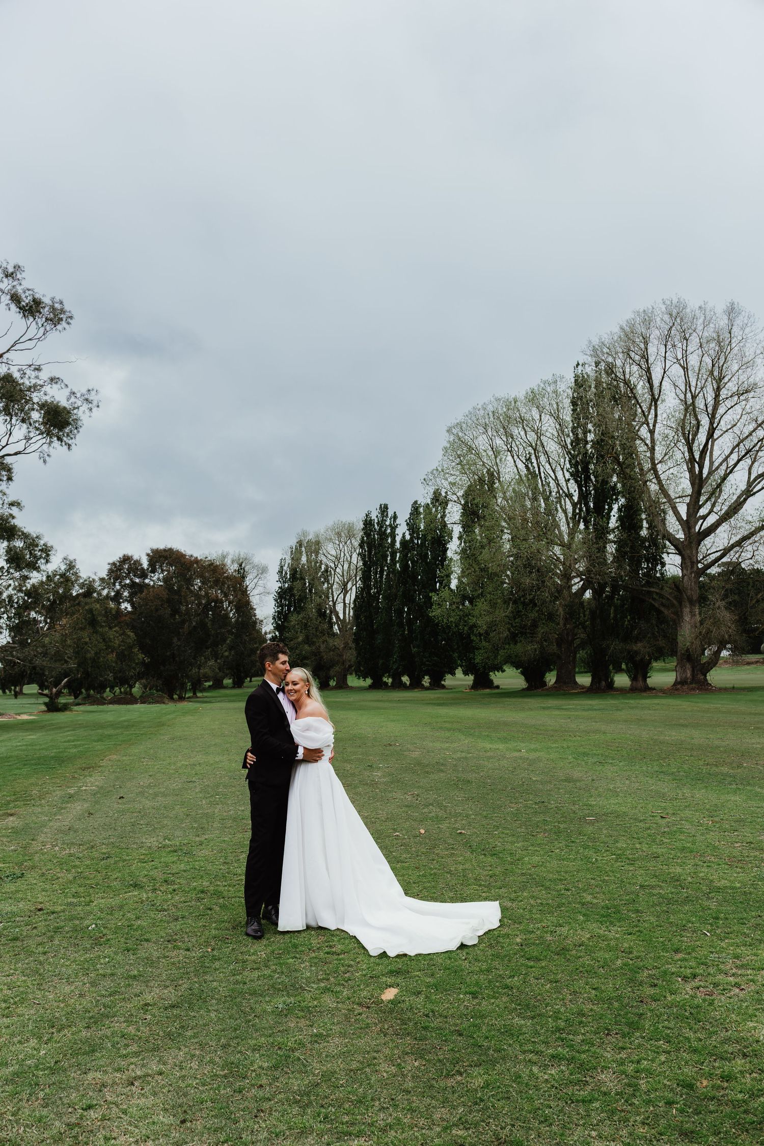 Bride and groom standing on lush green lawn surrounded by trees on their wedding day.