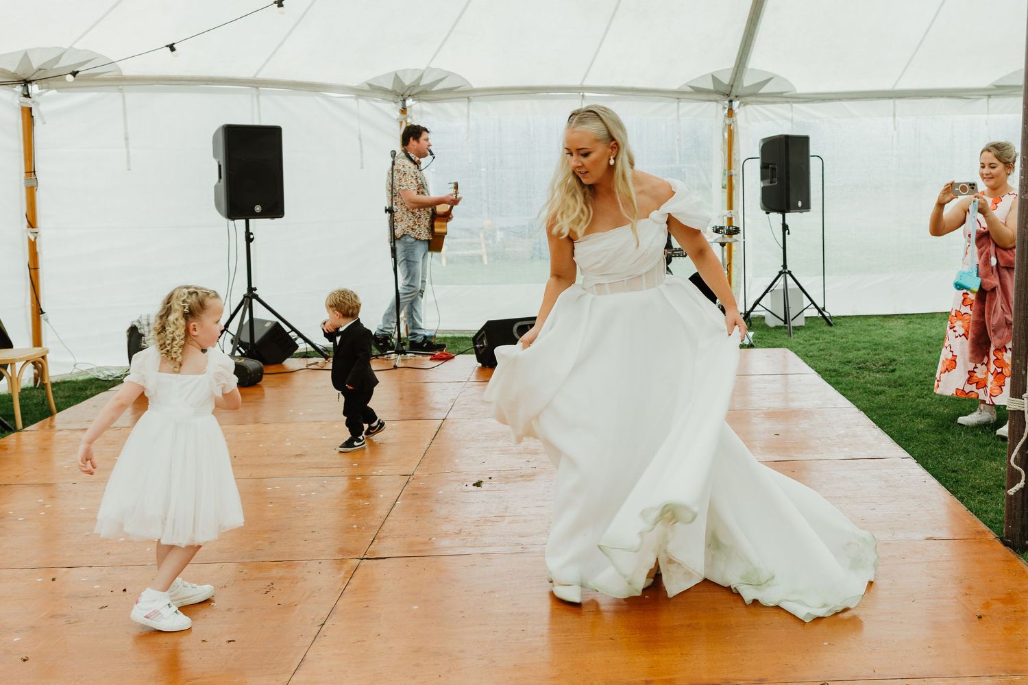 Bride in flowing white gown dances with young children on wooden reception dance floor.
