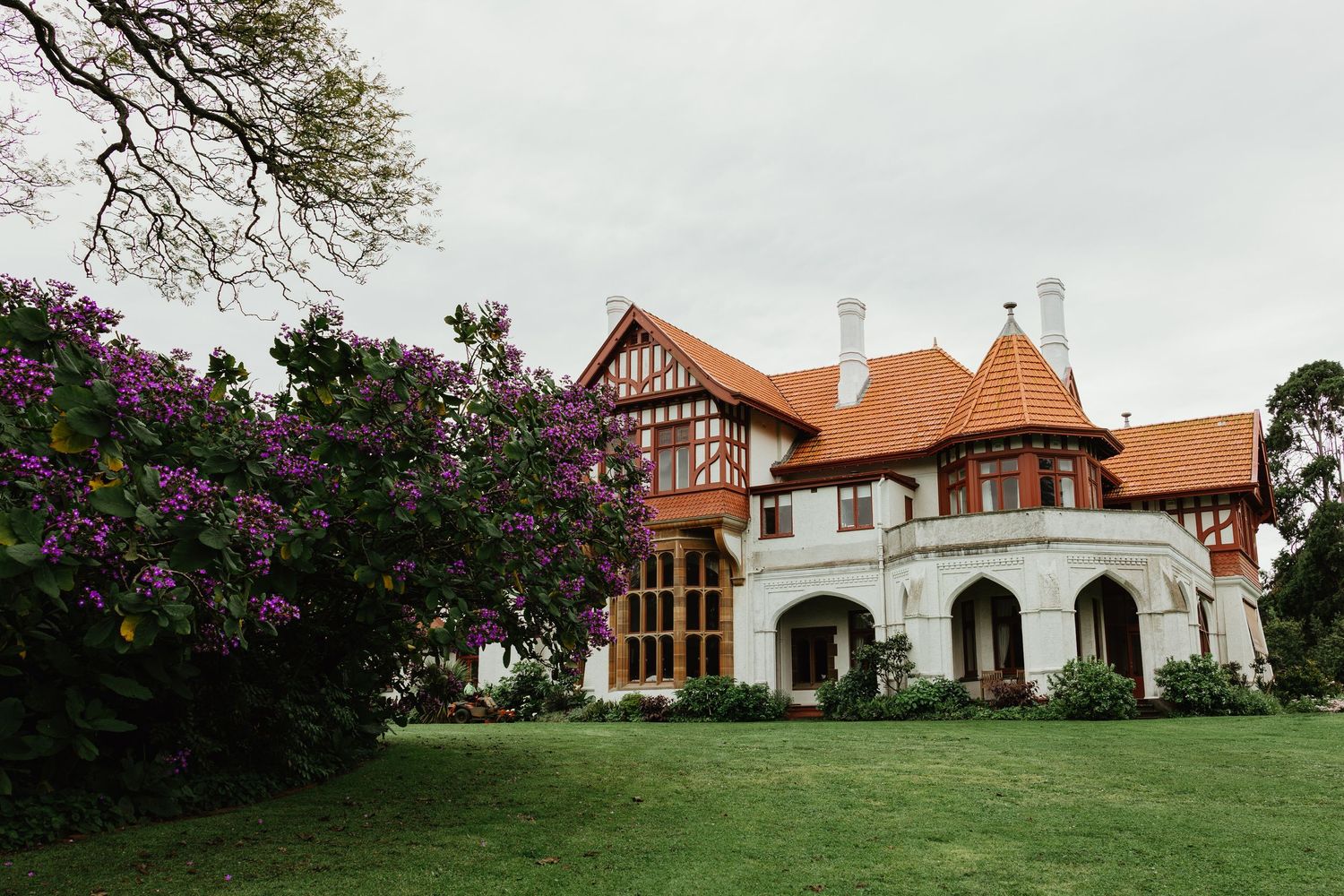 Stunning Tudor-style mansion with orange tiled roof surrounded by purple flowering bushes and manicured gardens.