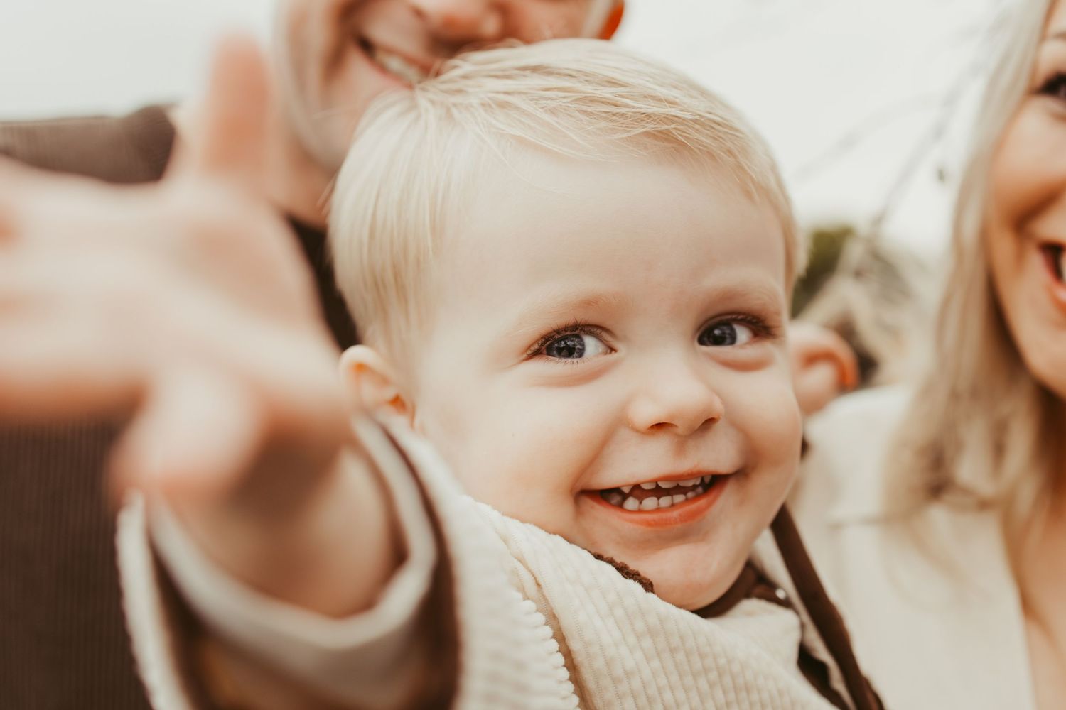 Smiling blonde toddler reaching toward camera during outdoor family photo session.