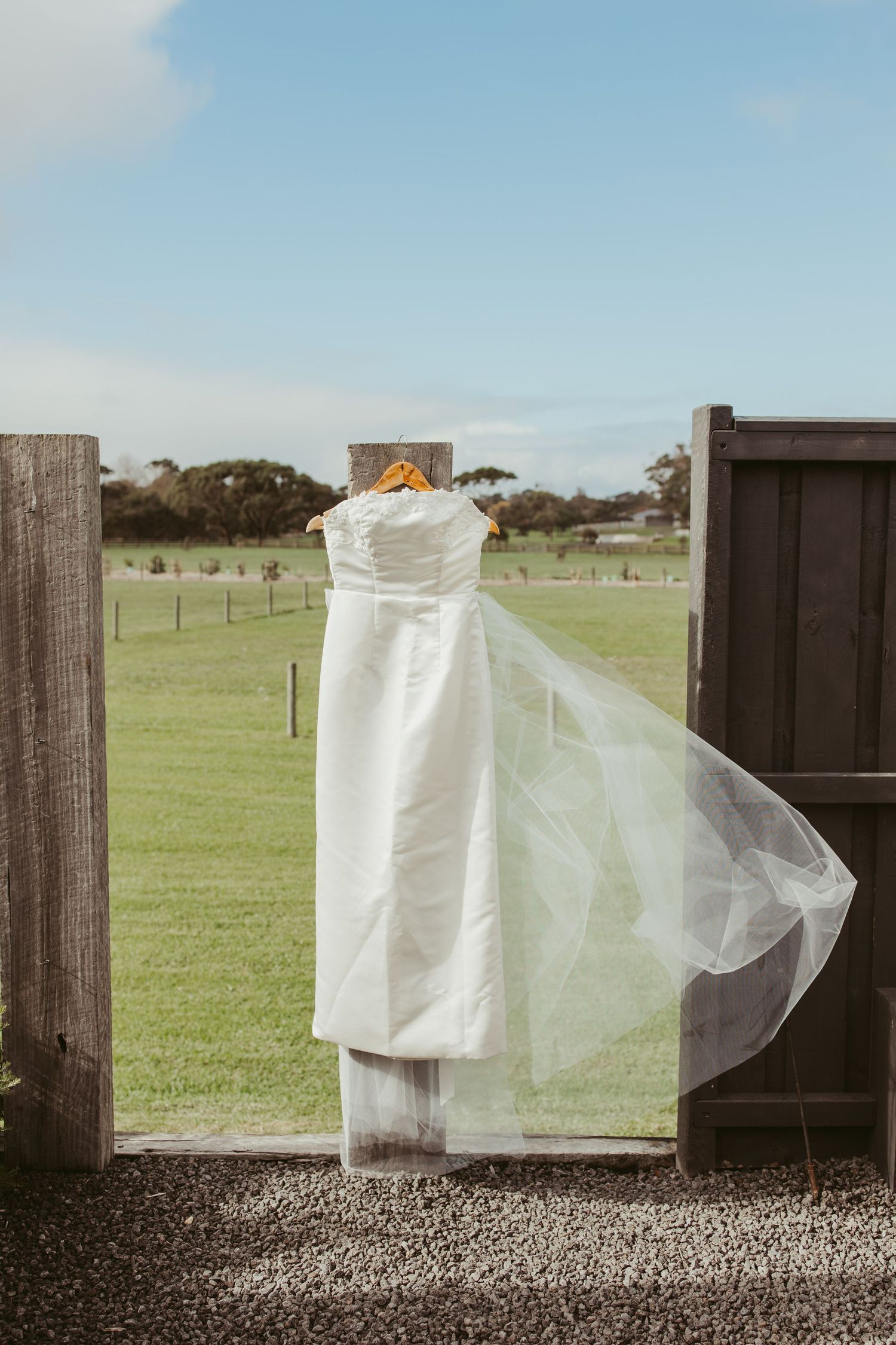 White wedding dress hanging on wooden hanger between rustic barn doors with green countryside backdrop.