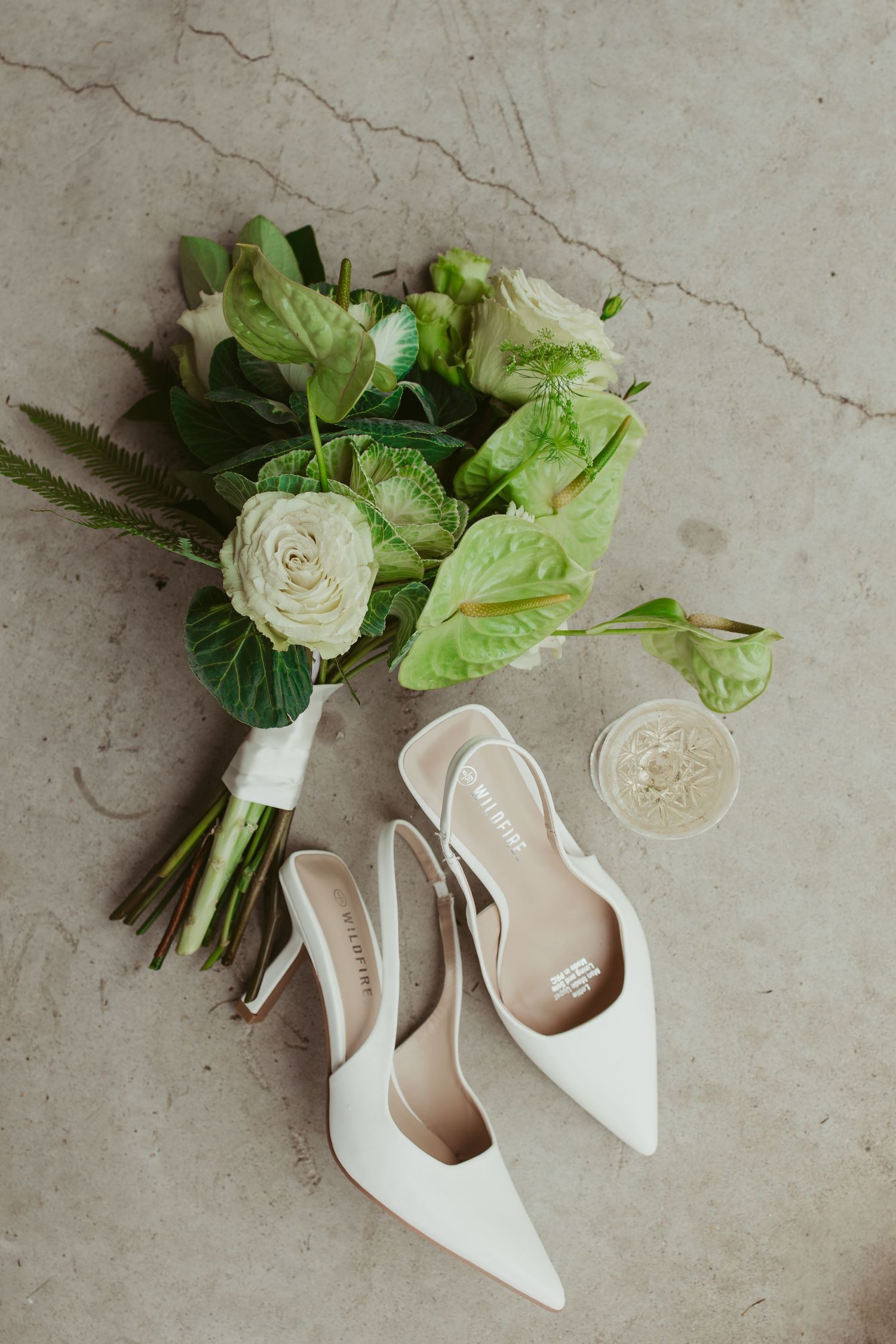 White bridal heels, green floral bouquet with roses, and a ring dish arranged on a marble surface.