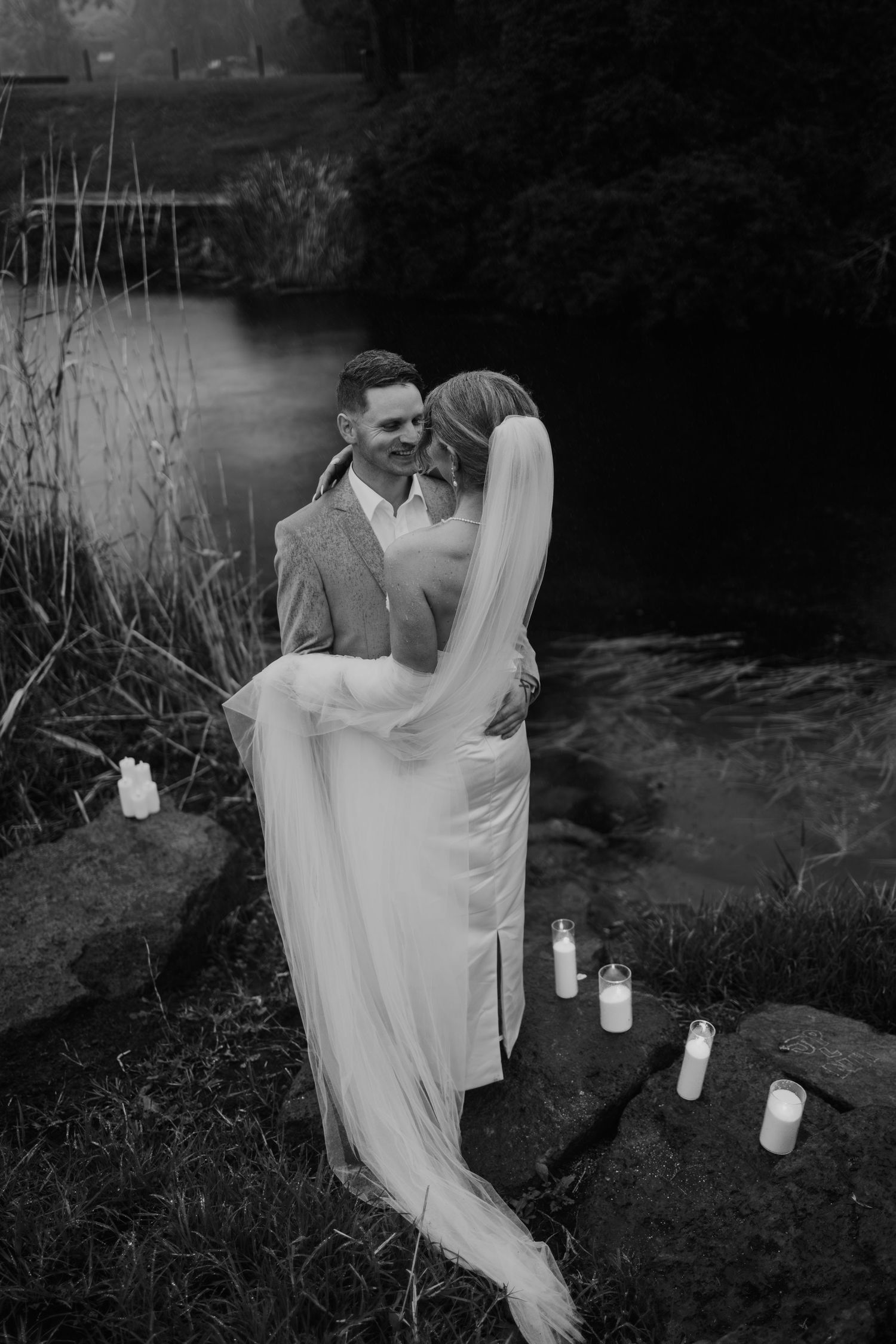 Black and white photo of a groom lifting his bride near a candlelit riverside setting on their wedding day.