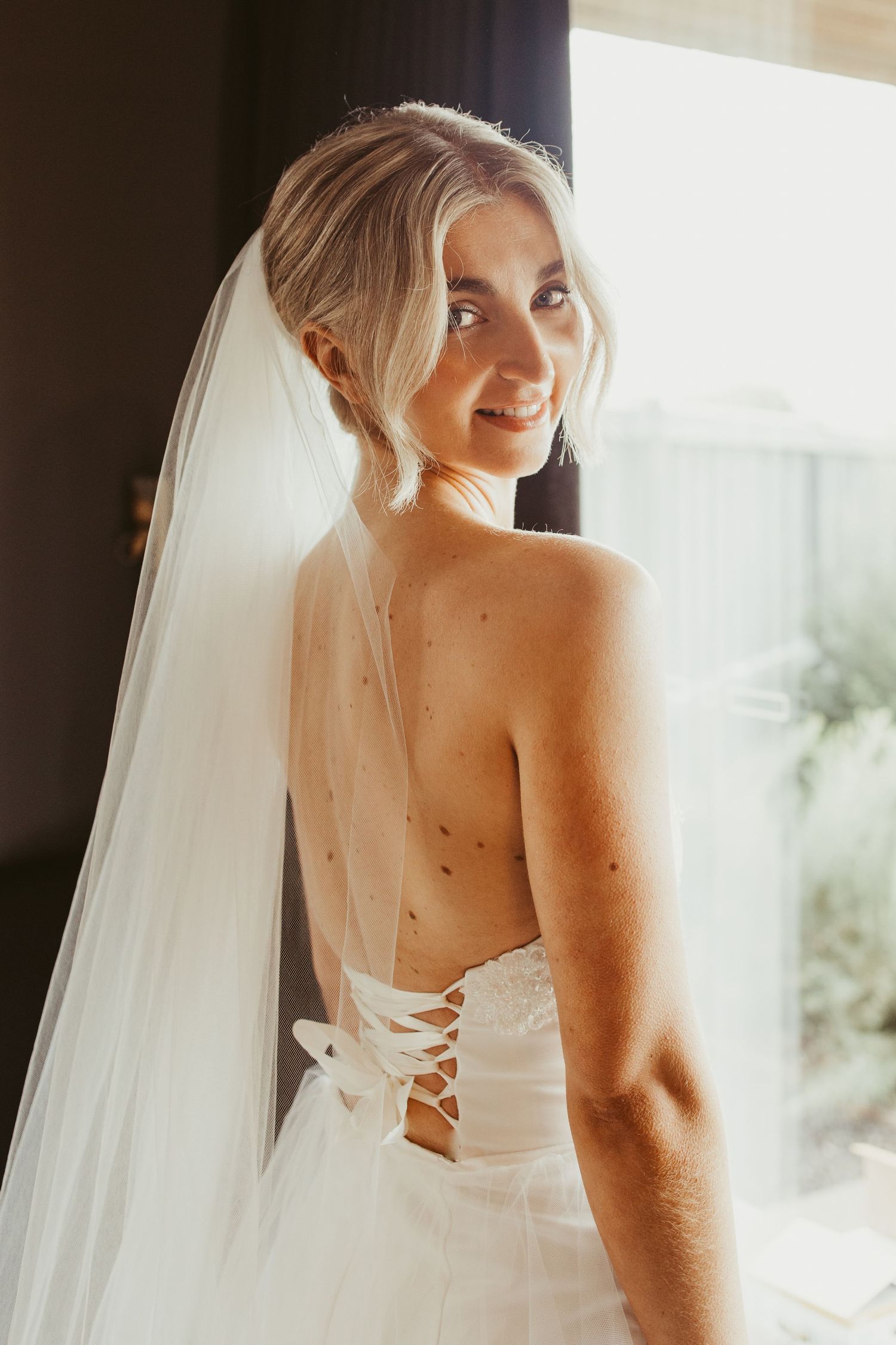 Blonde bride in backless lace-up wedding gown and veil smiling over shoulder near window.