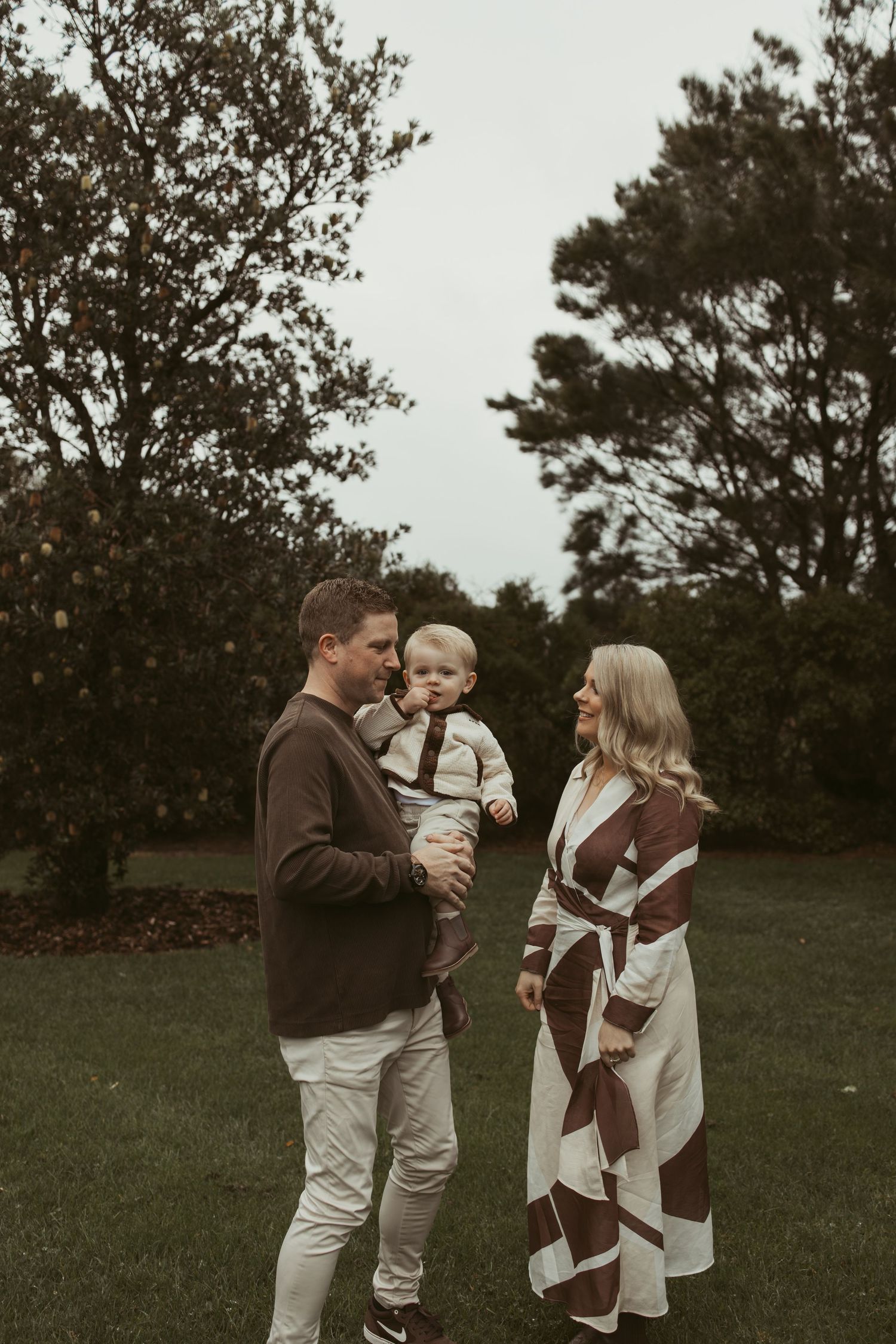 Family of three with baby in autumn park, father in brown sweater, mother in brown striped coat.