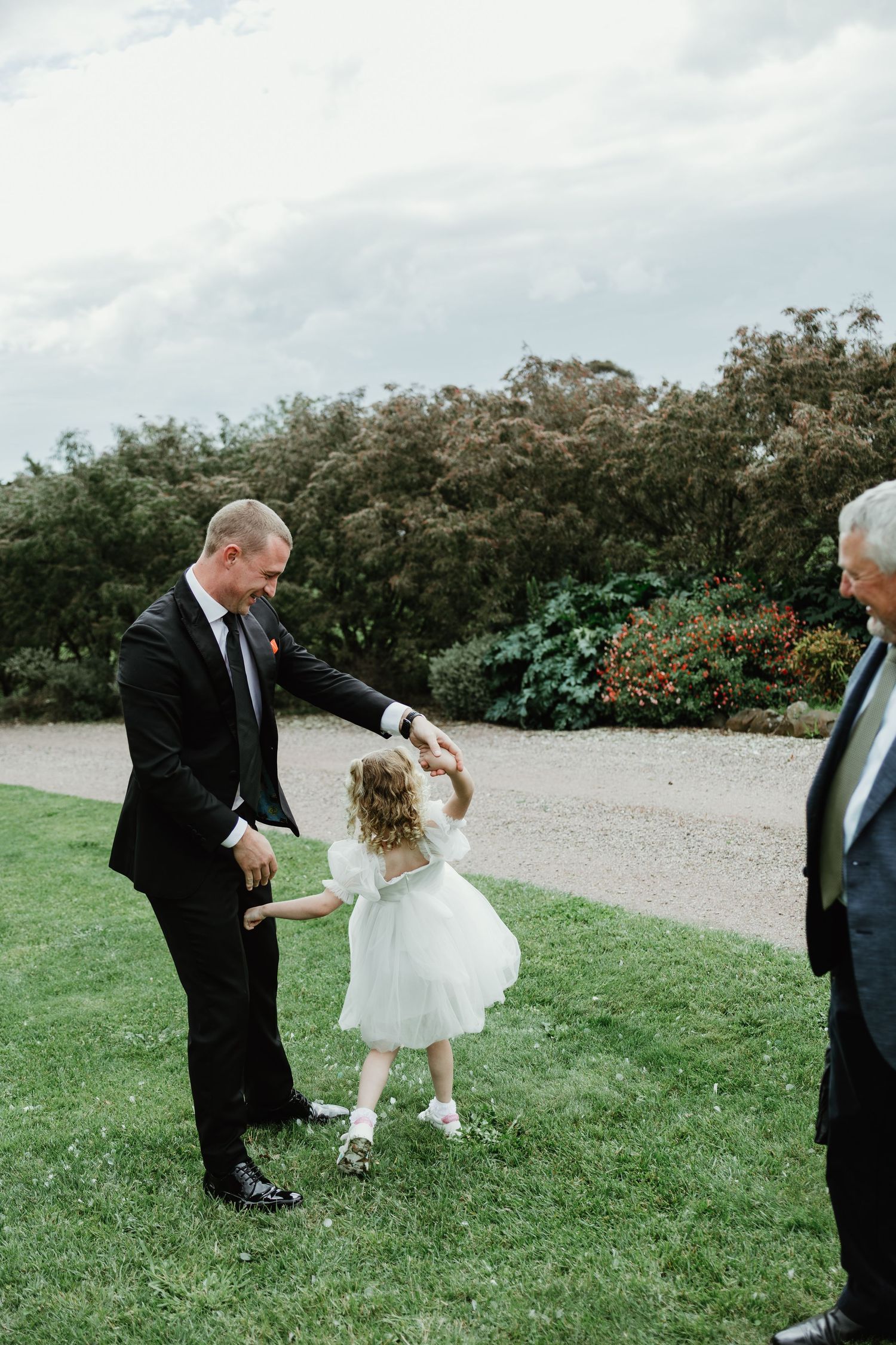 Man in black suit twirls young flower girl in white dress on wedding venue lawn.