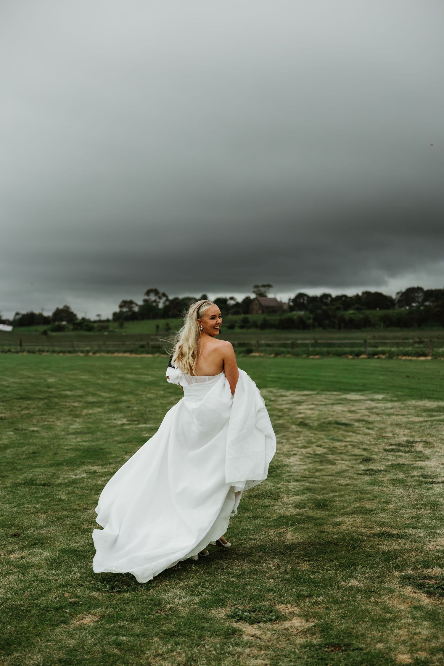 Bride in flowing white gown twirling in open field under dramatic stormy grey sky.
