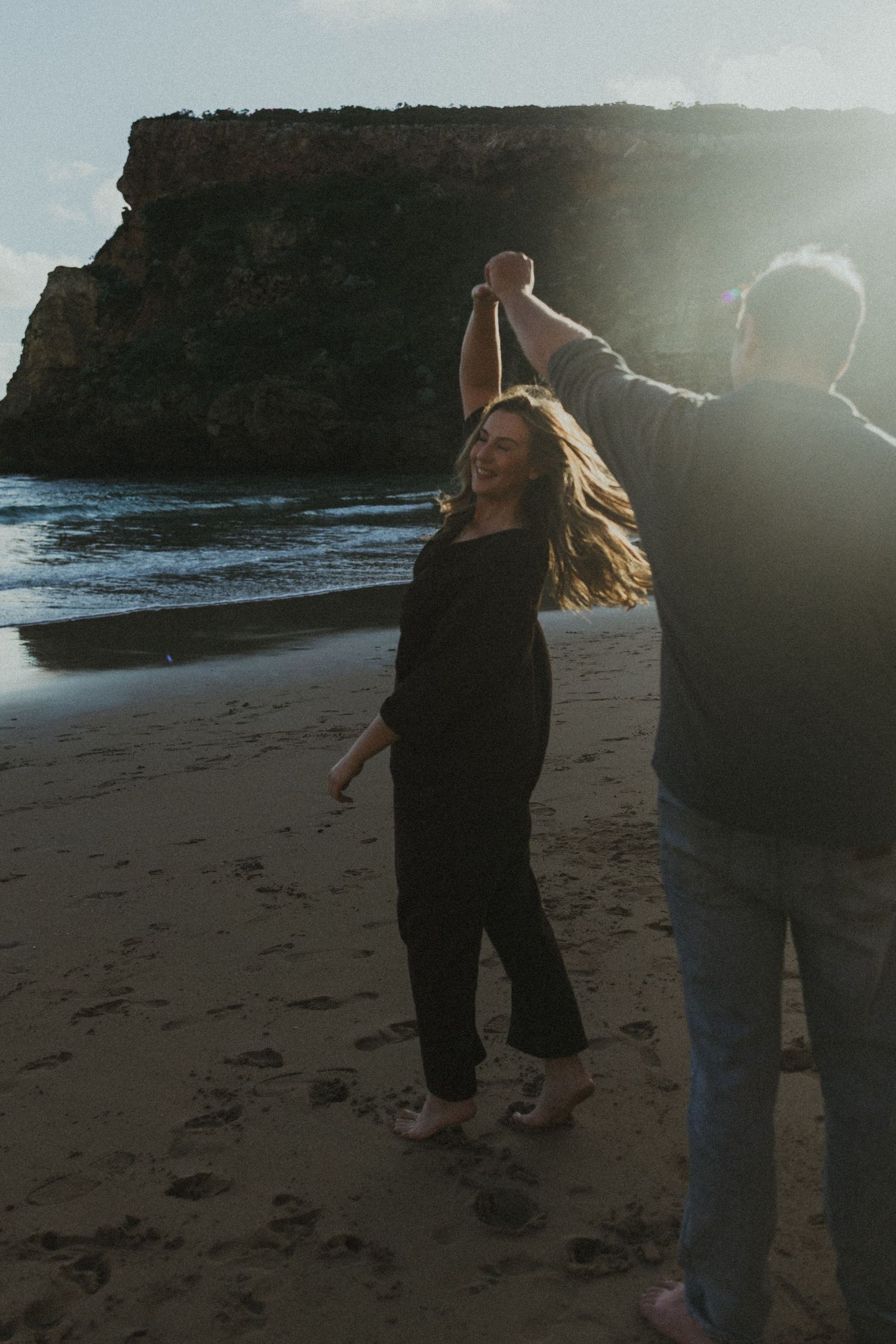 A man spins a woman by her hand on a beach at sunset with rocky cliffs in the background.