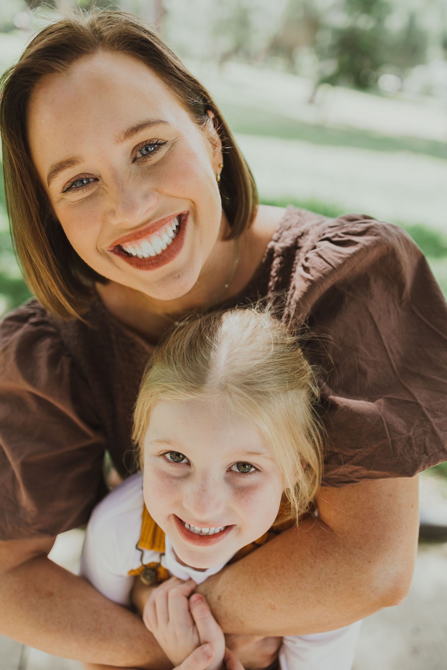 A smiling mother hugs her young blonde daughter from behind in a sunny park setting.