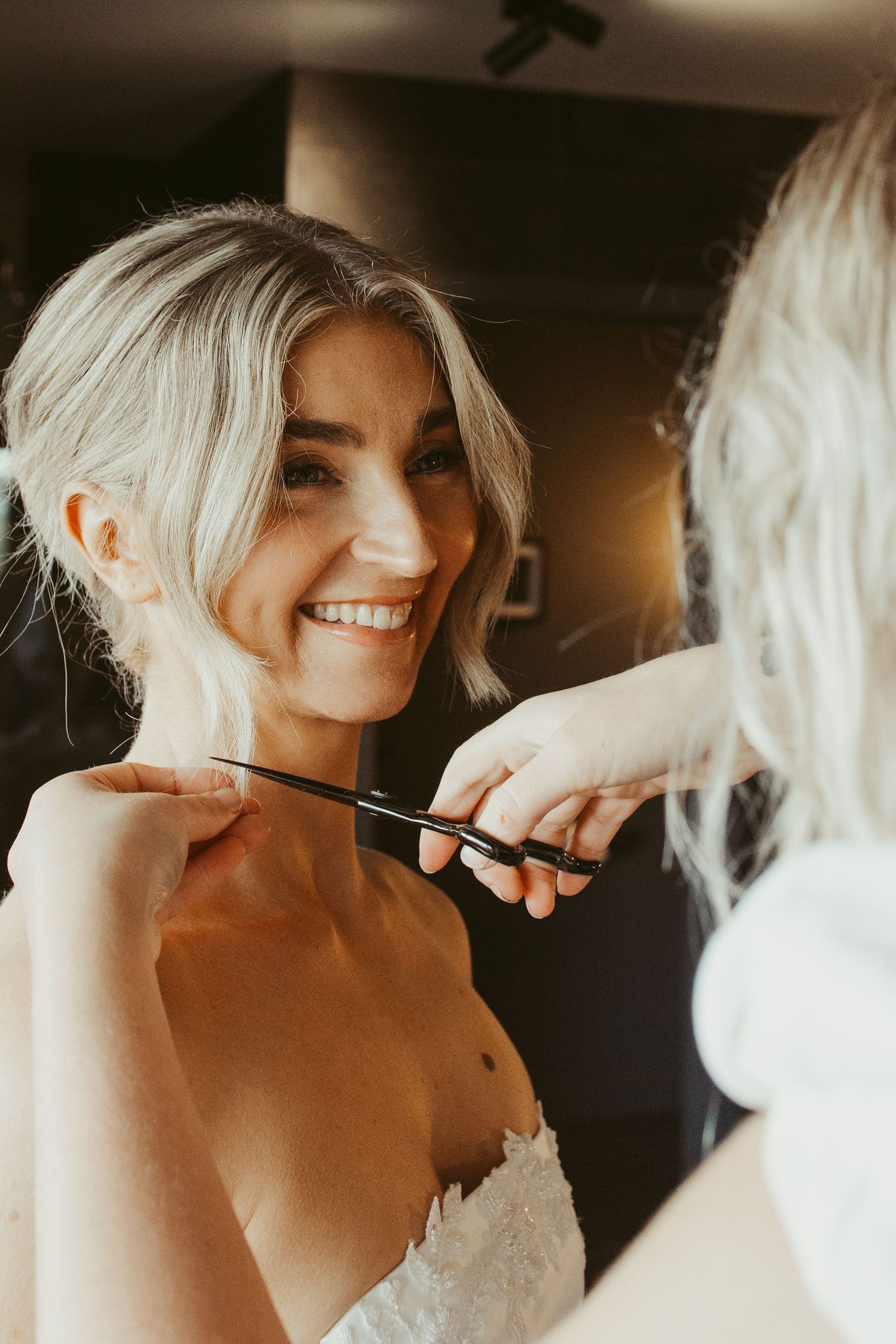 Smiling bride in strapless wedding dress getting finishing touches done by makeup artist in mirror.