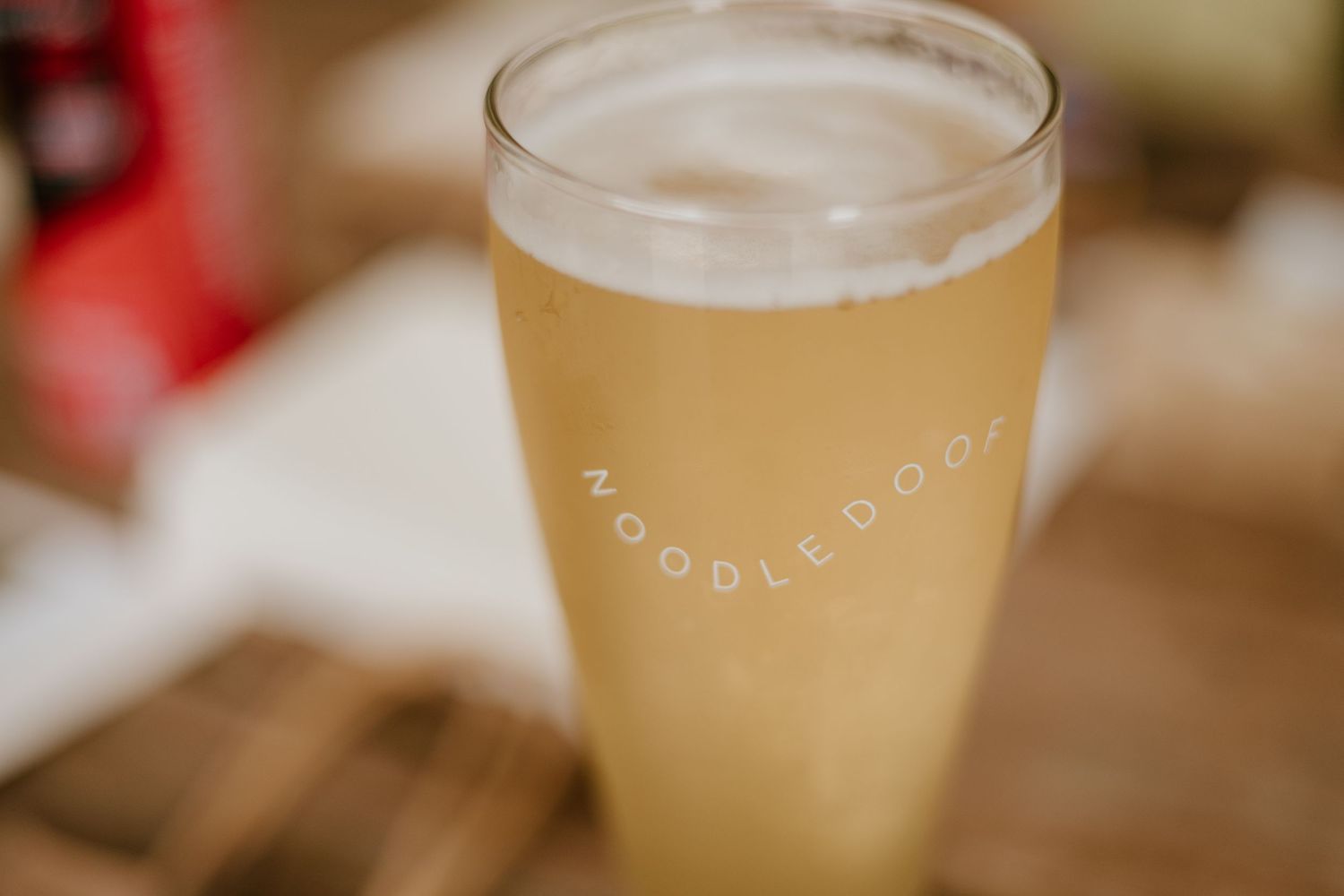 A tall Noodledoor branded pint glass filled with golden beer on a wooden surface.