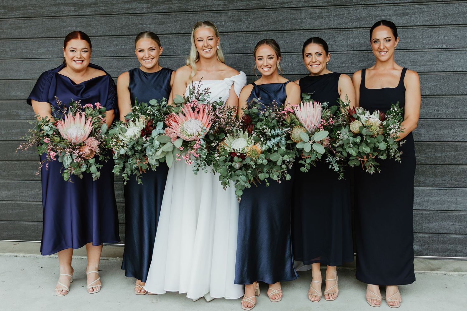 Bride in white gown with five bridesmaids in navy dresses holding large protea and greenery bouquets.