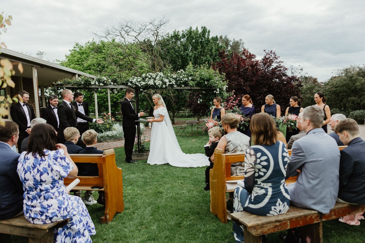 Outdoor wedding ceremony with guests seated on wooden pews as couple exchanges vows under floral arch.