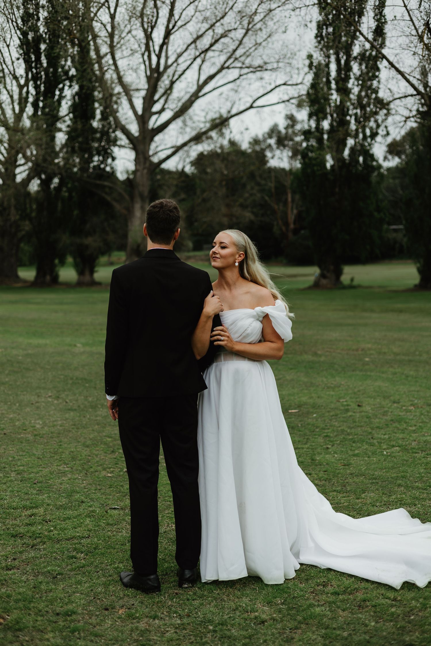 Bride in elegant off-shoulder white gown smiling at groom in black suit on lawn.