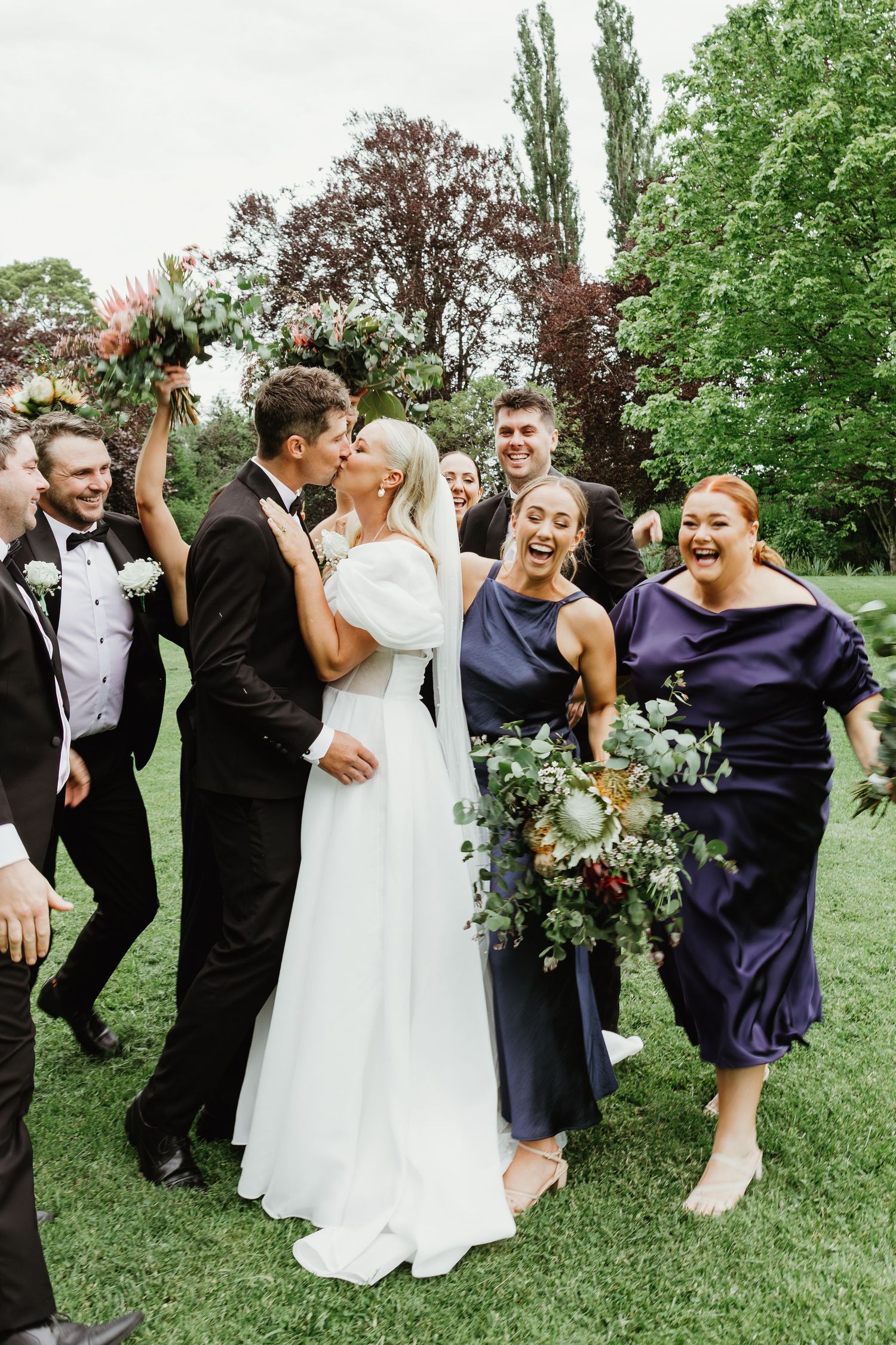 Bride and groom share a kiss surrounded by laughing wedding party in navy attire on a lush green lawn.