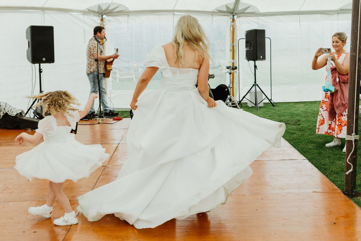 Bride in off-shoulder gown twirls with flower girl on dance floor while live band performs.