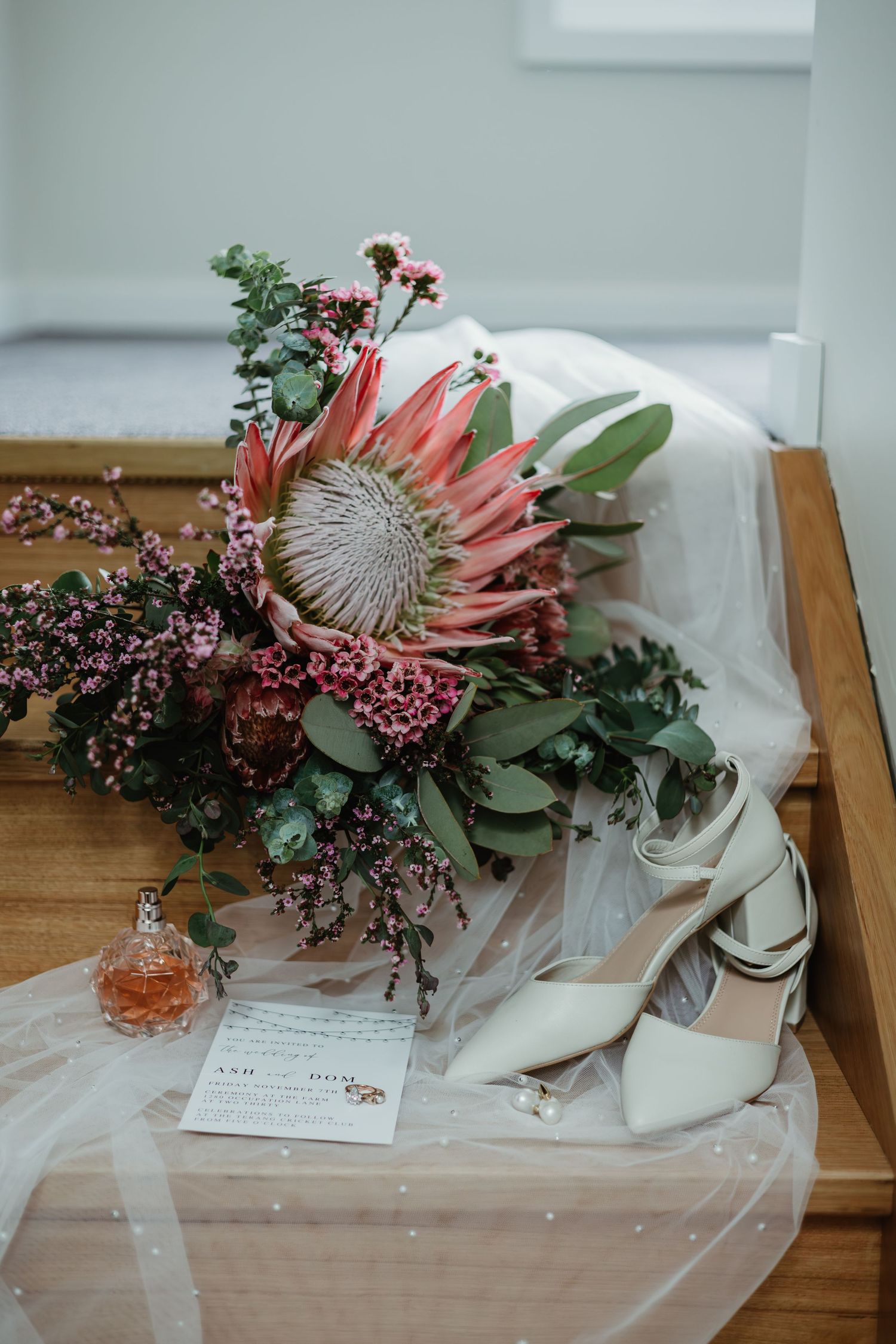 Bridal flat lay with king protea bouquet, white heels, perfume, and veil on wooden bench.