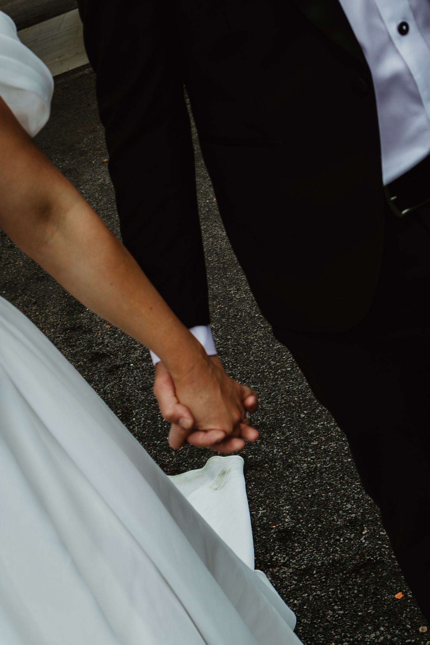 Bride and groom holding hands, bride in white gown and groom in black suit, close-up detail shot.