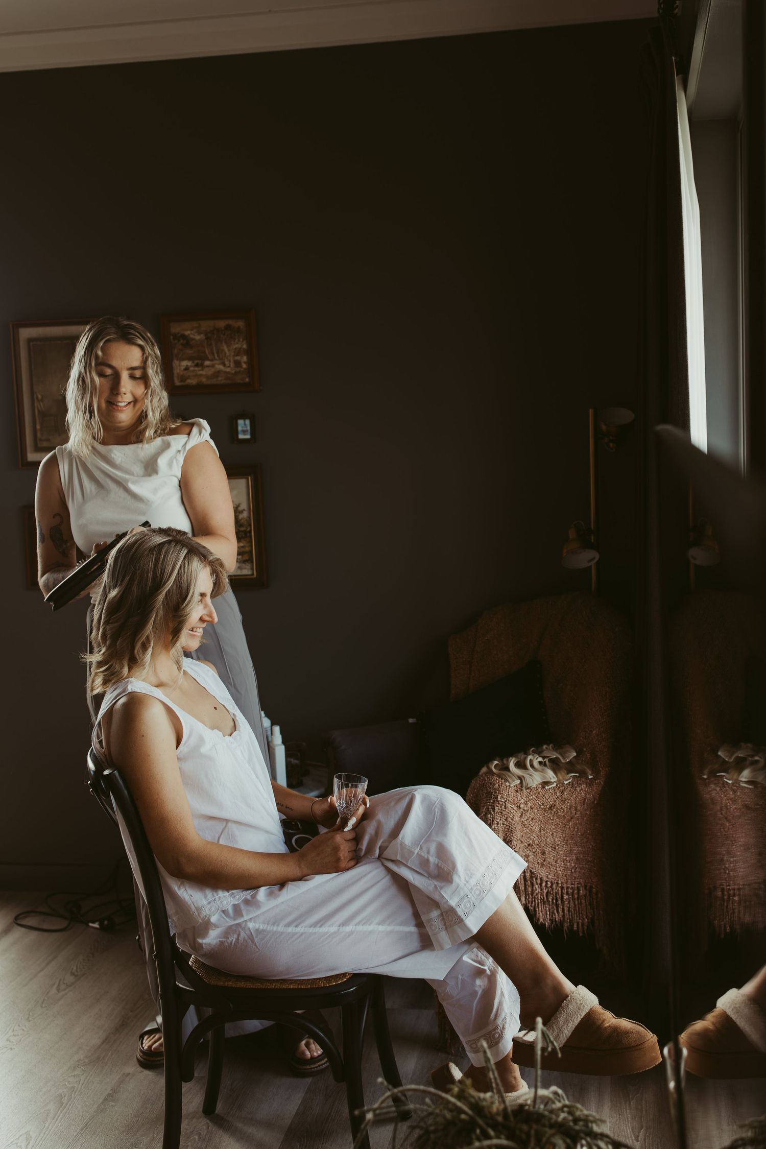 Two blonde bridesmaids in white getting ready together in a dark moody bedroom before a wedding.