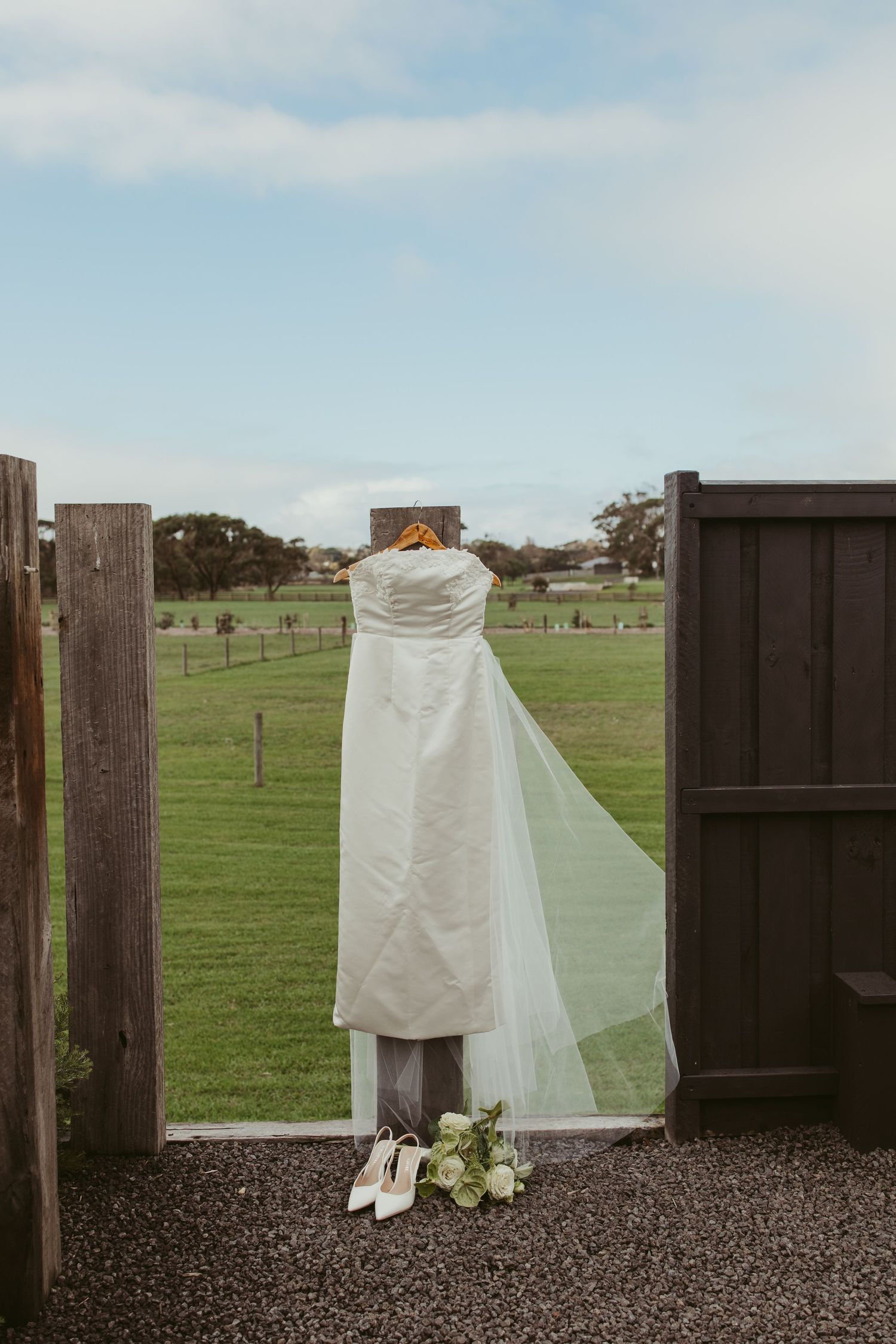 A strapless white wedding dress with veil hangs between barn doors overlooking a green field.