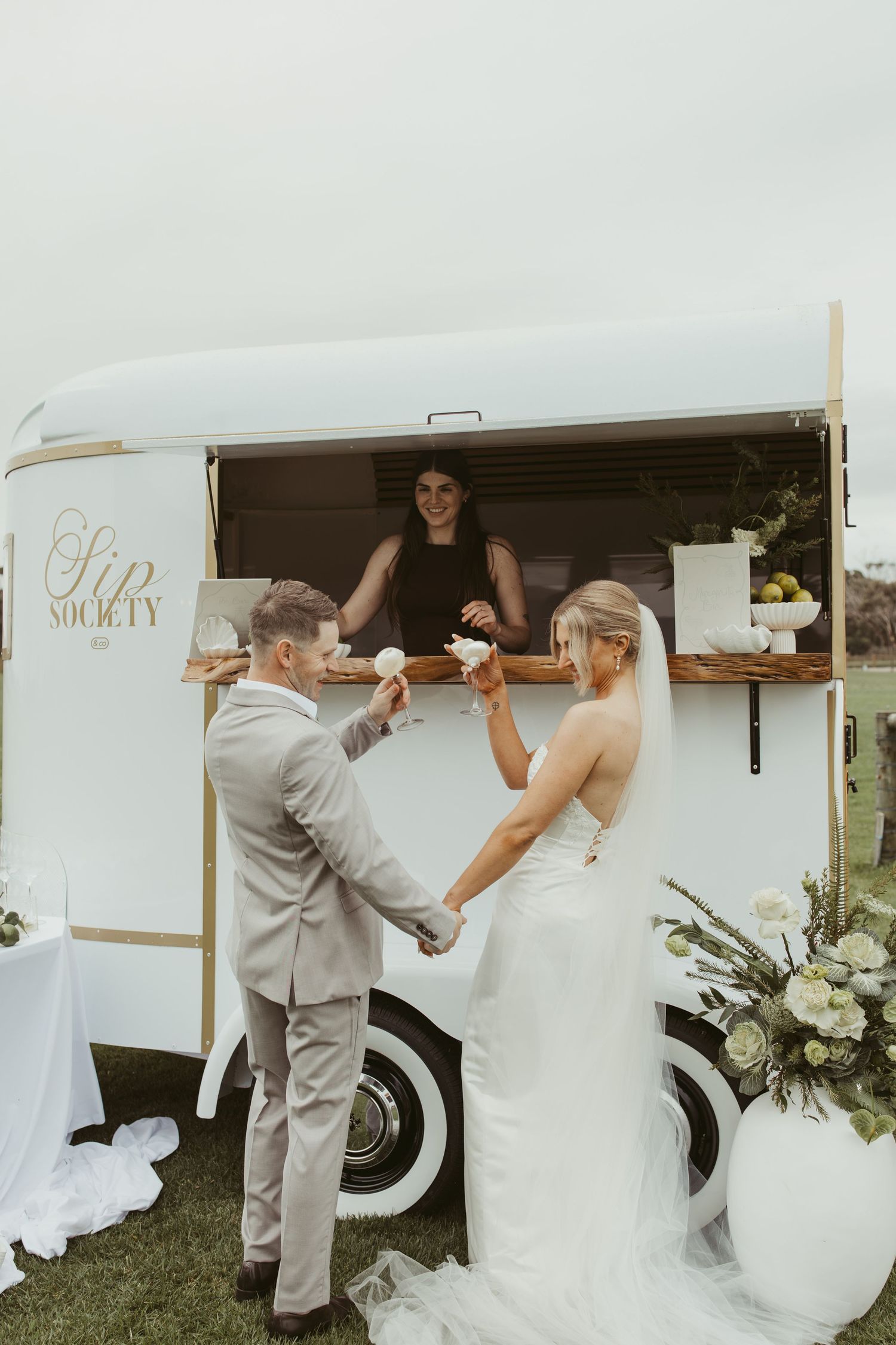 Newlyweds toasting with drinks served from a vintage white Sip Society mobile bar at their wedding.