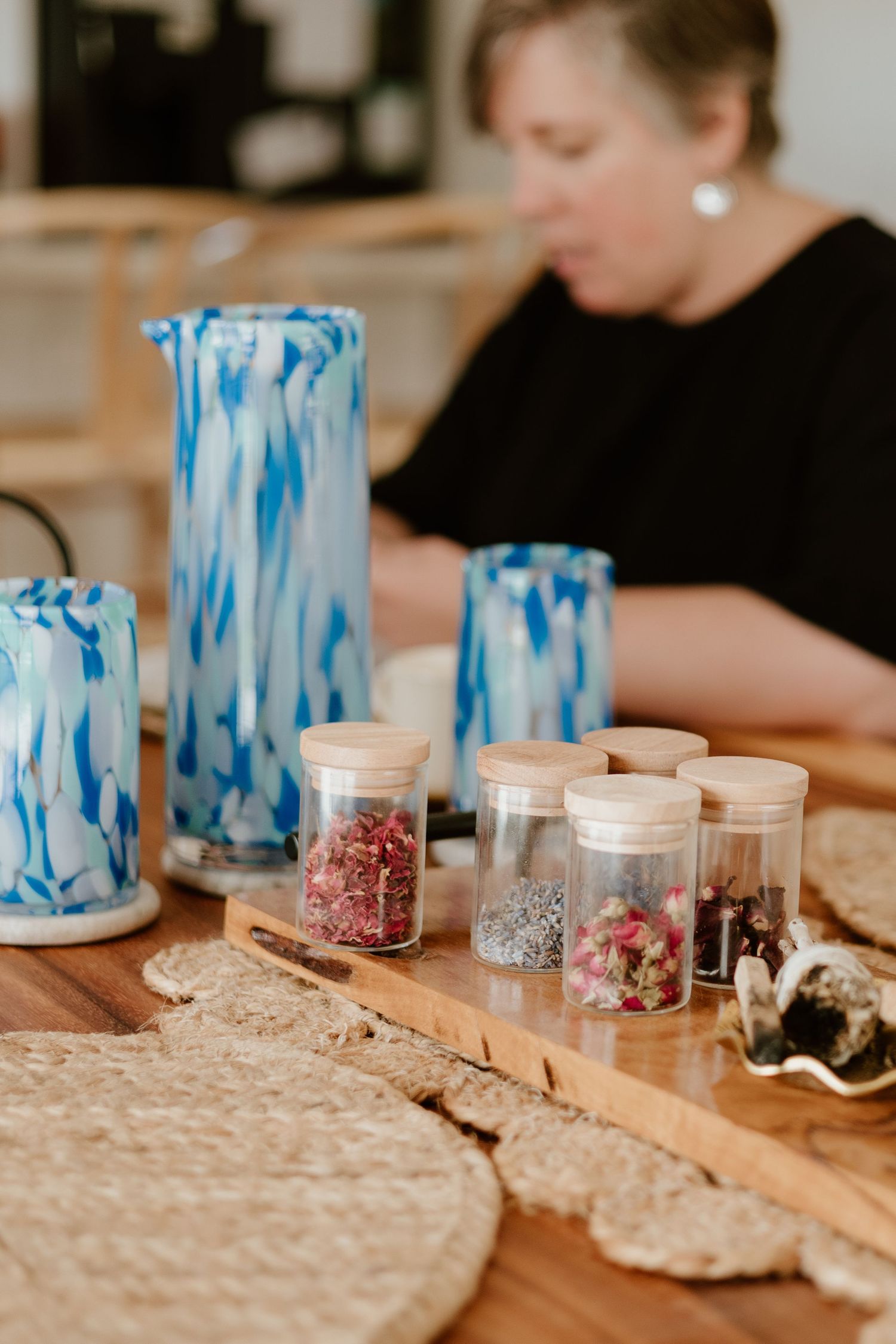 Blue marbled glass pitcher and tumblers with small spice jars on wooden board, woman in black blouse working in background.
