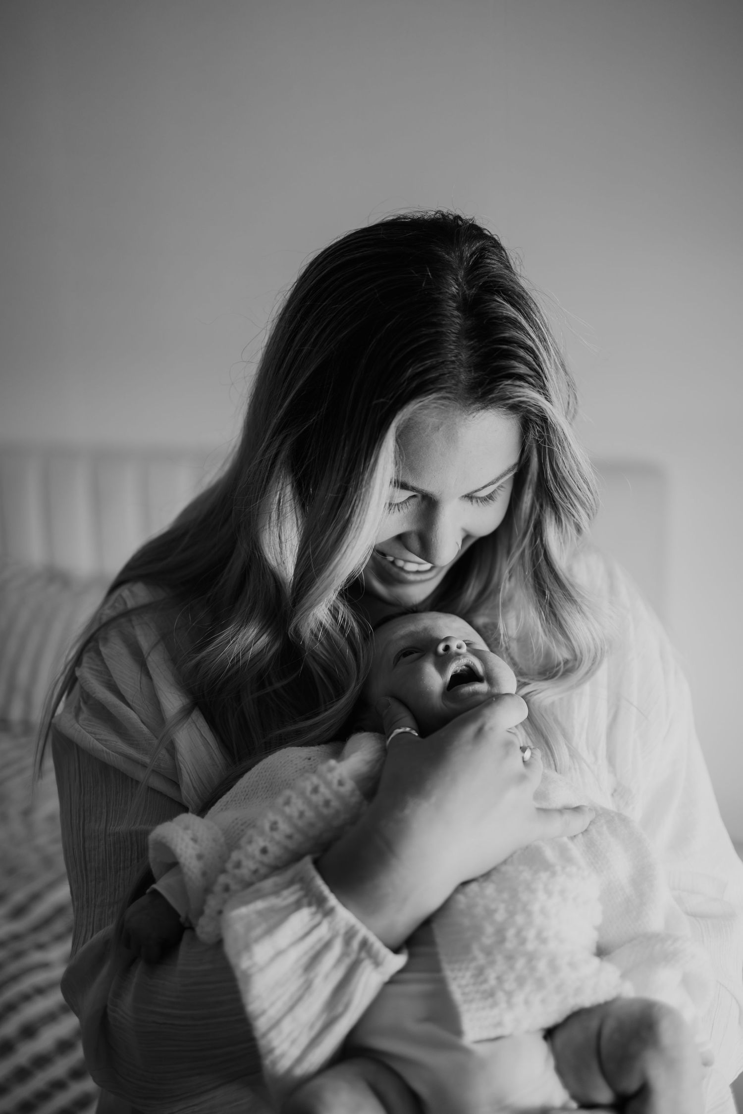 Black and white photo of a smiling mother tenderly holding her yawning newborn baby while sitting on a bed.