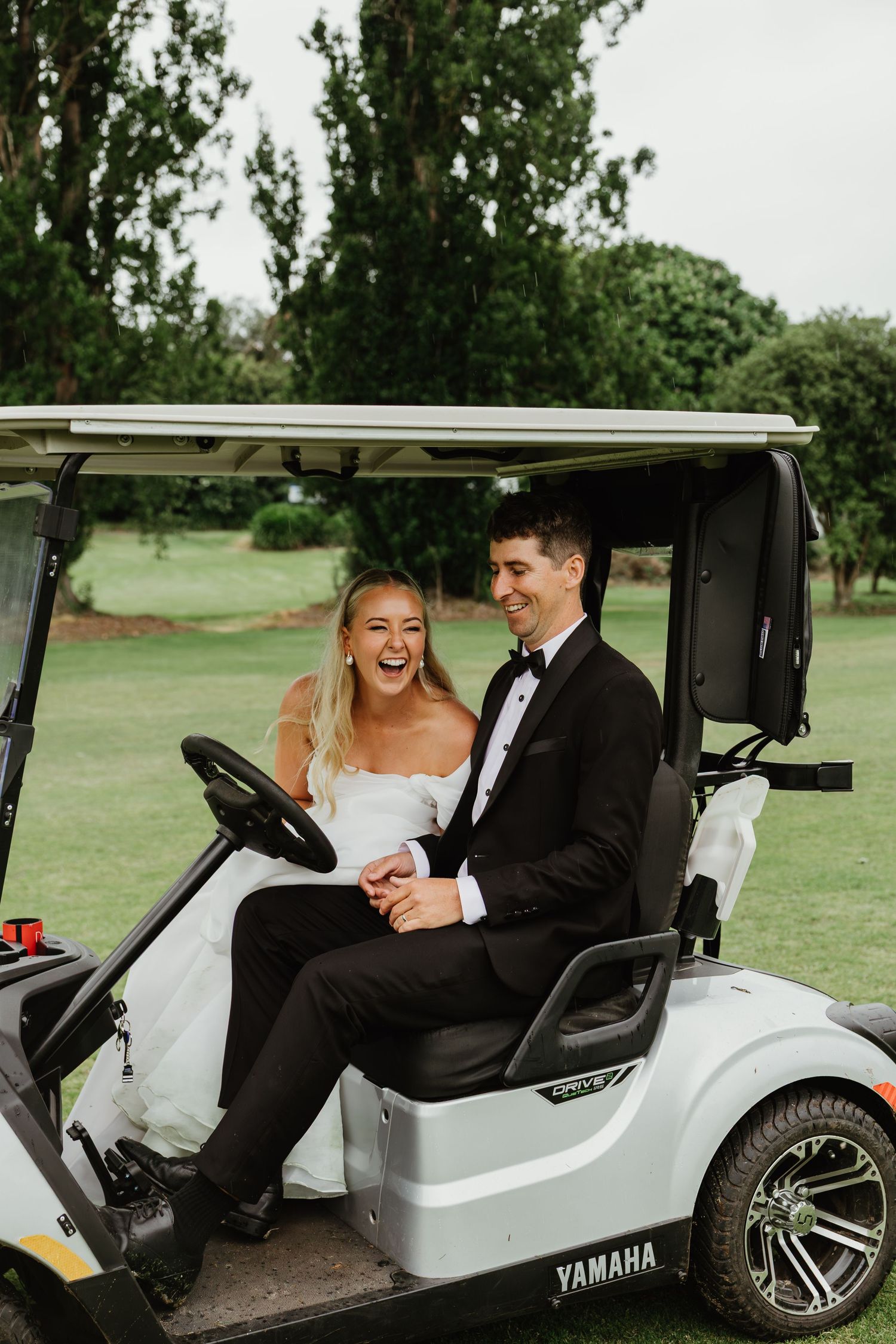 Laughing bride and groom in wedding attire ride a Yamaha golf cart on a lush green course.