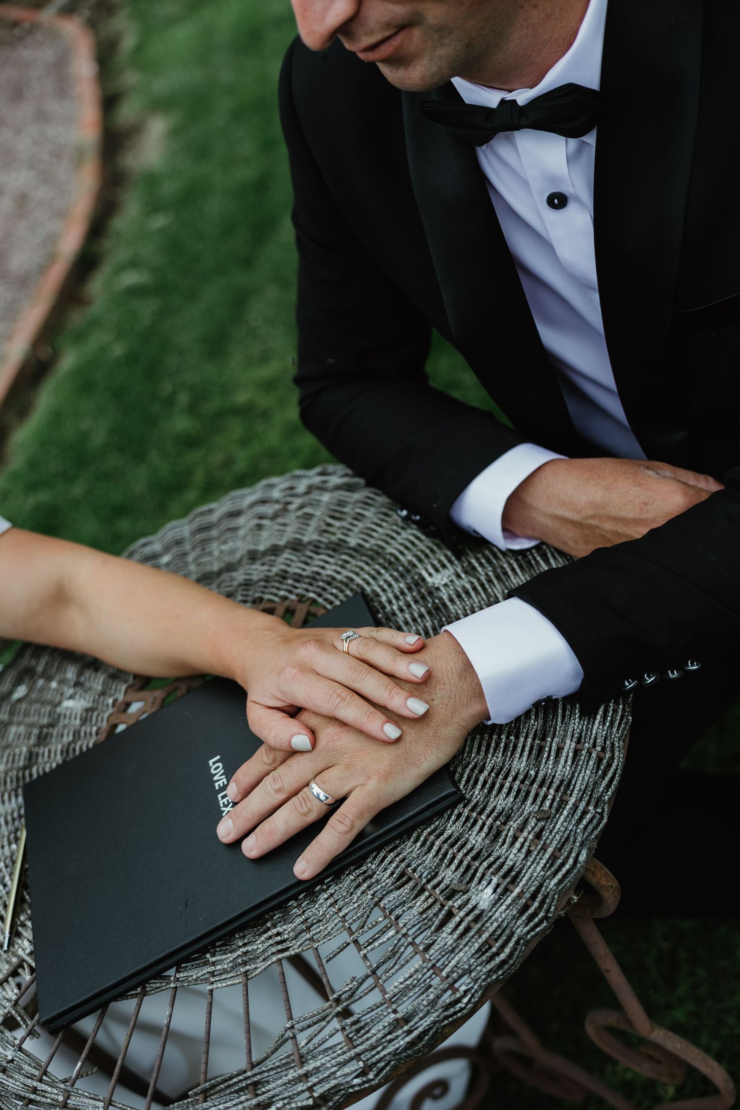 Bride and groom holding hands over a wedding album on a wicker chair, showcasing elegant rings and formal attire.