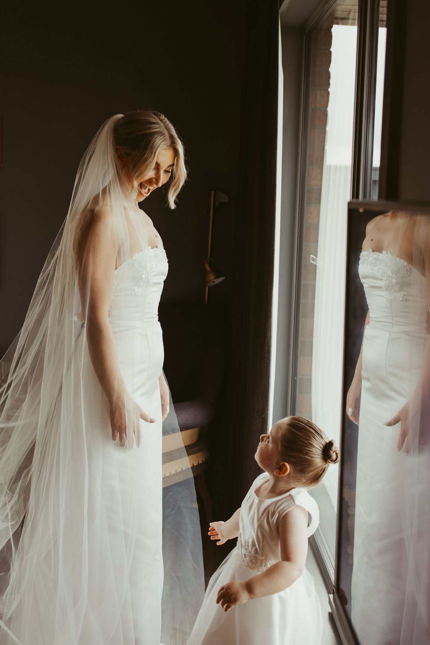 Smiling bride in white strapless gown and veil interacting with young flower girl by a sunlit window.