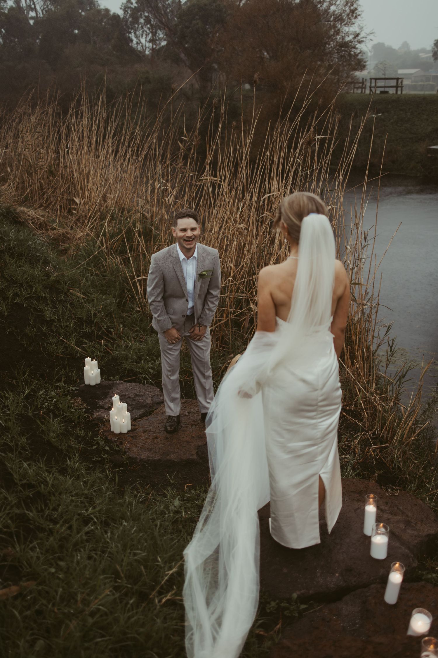 Bride and groom share an intimate first look ceremony by a candlelit waterside setting surrounded by tall reeds.