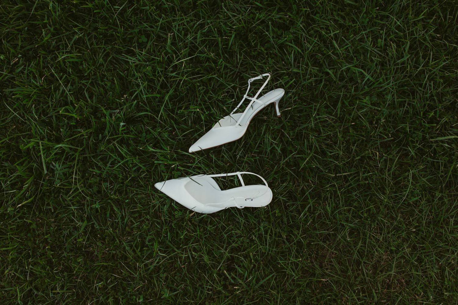 Pair of white slingback heeled shoes lying on lush green grass.