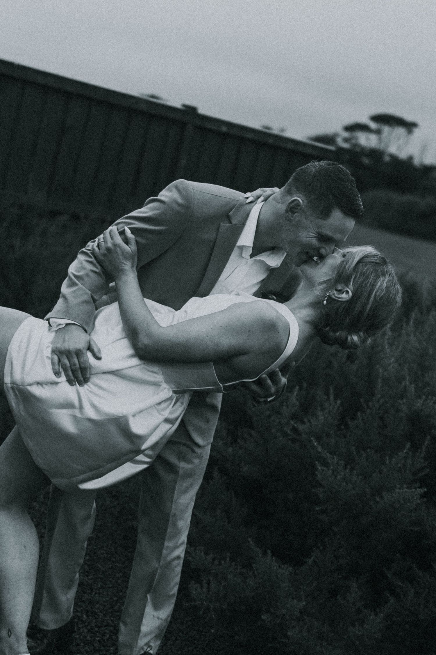 Groom dipping bride for a kiss outdoors in a dramatic black and white wedding portrait.