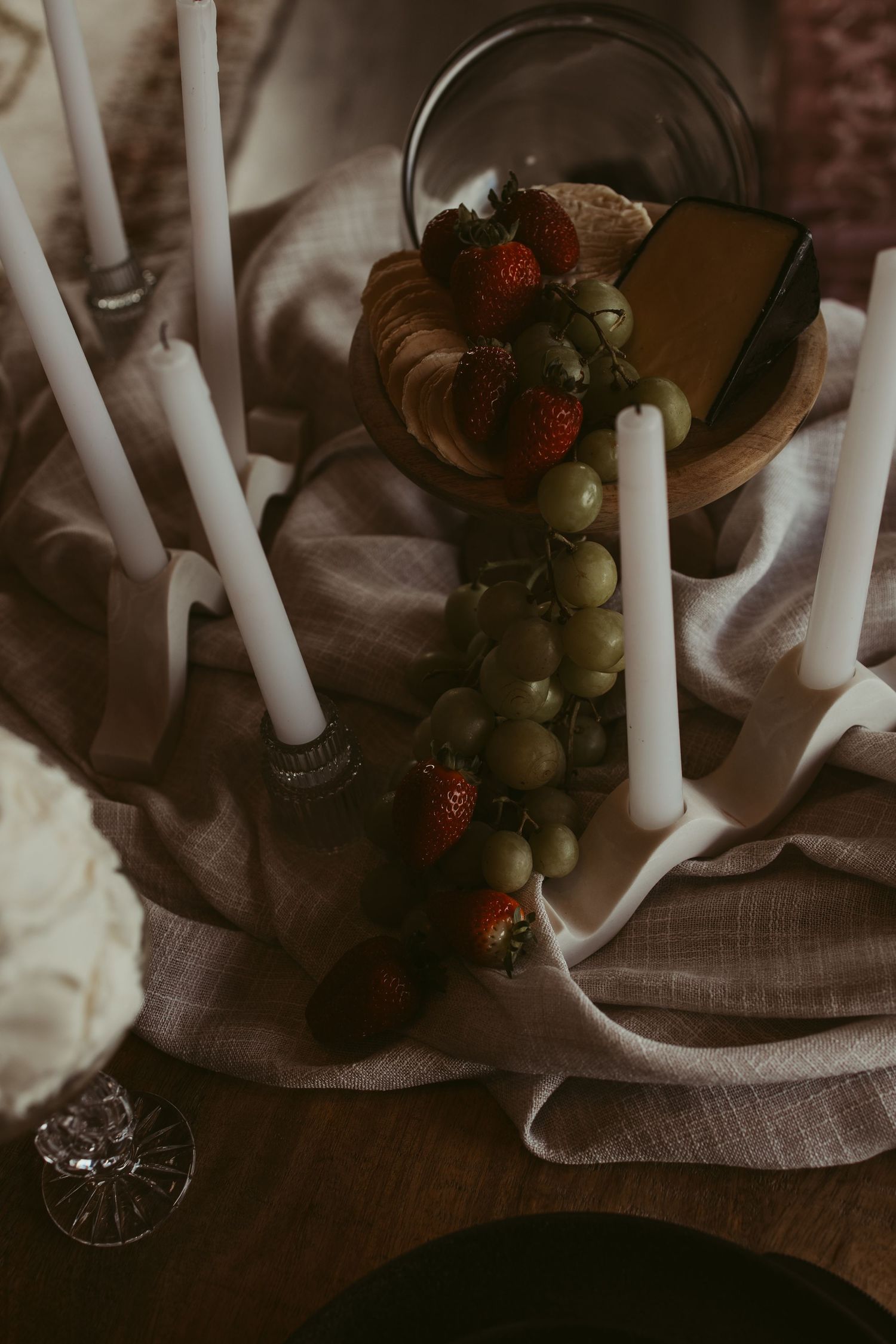 Elegant wedding tablescape with white taper candles, fresh strawberries, green grapes, and cheese on linen cloth.
