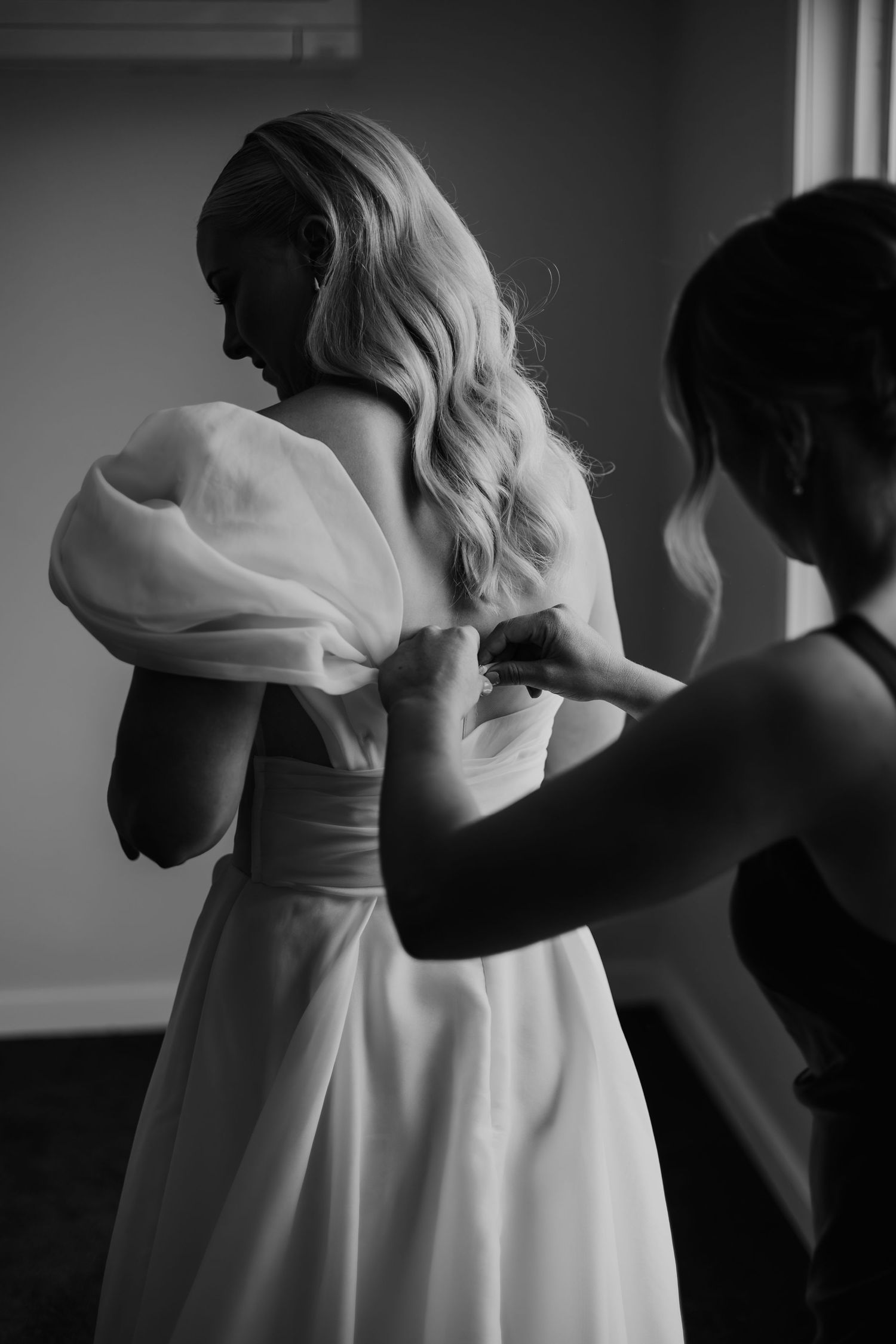 Bridesmaid fastening buttons on back of brides white puff sleeve wedding gown in black and white photo.