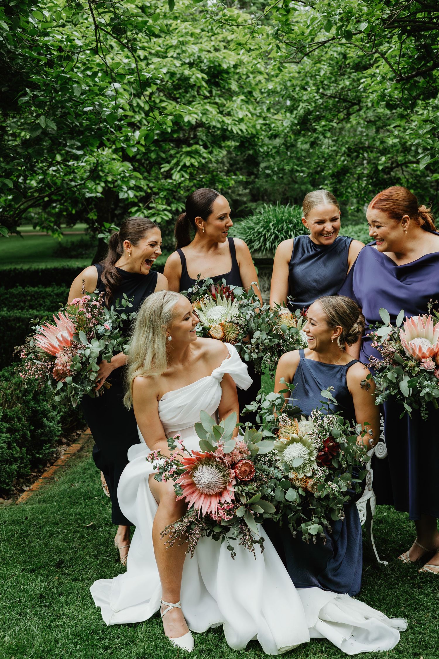Bride seated on grass with bridesmaids holding large native protea and greenery bouquets.