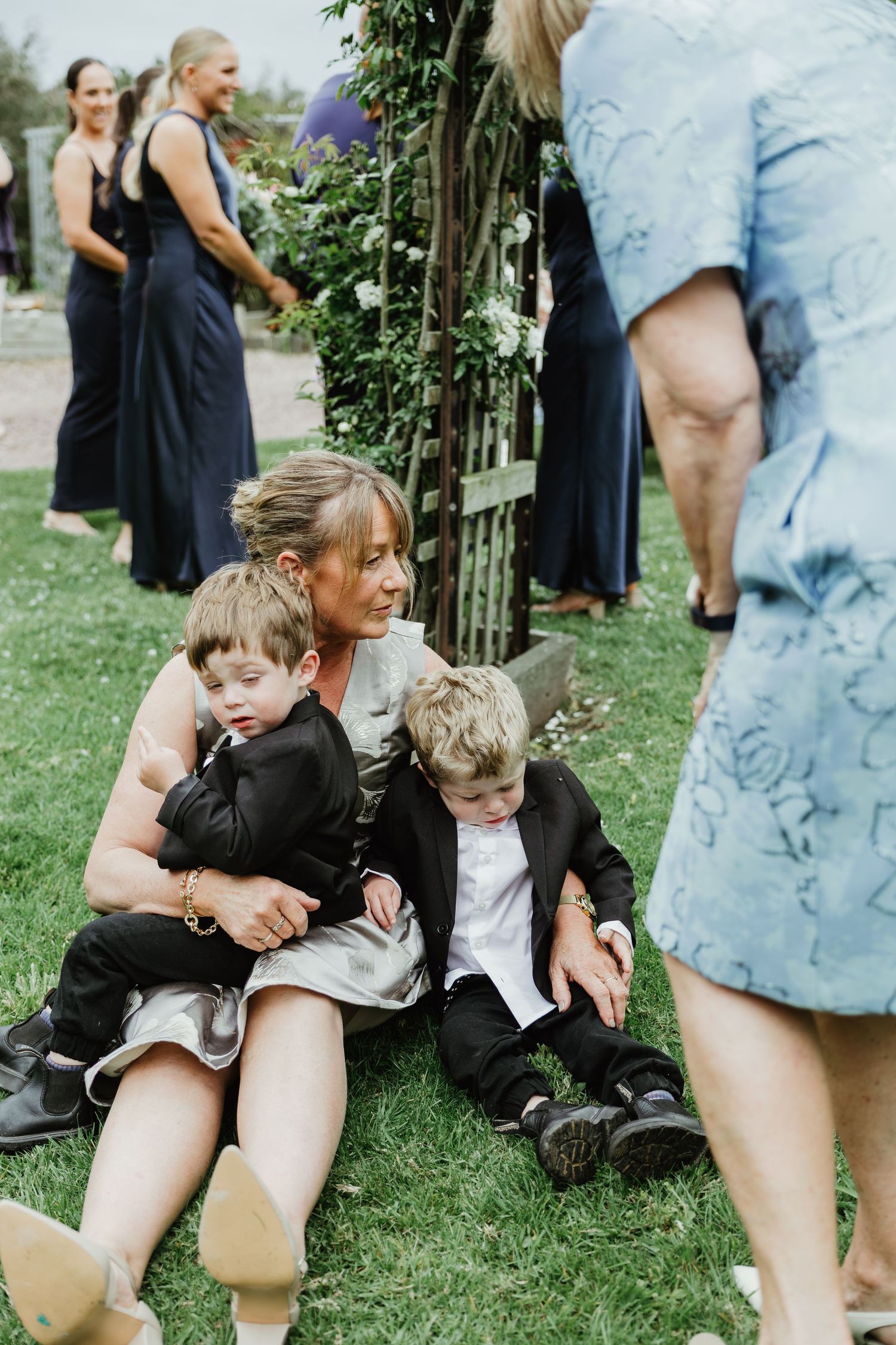 Woman sitting on grass with two young boys in suits during outdoor wedding celebration.