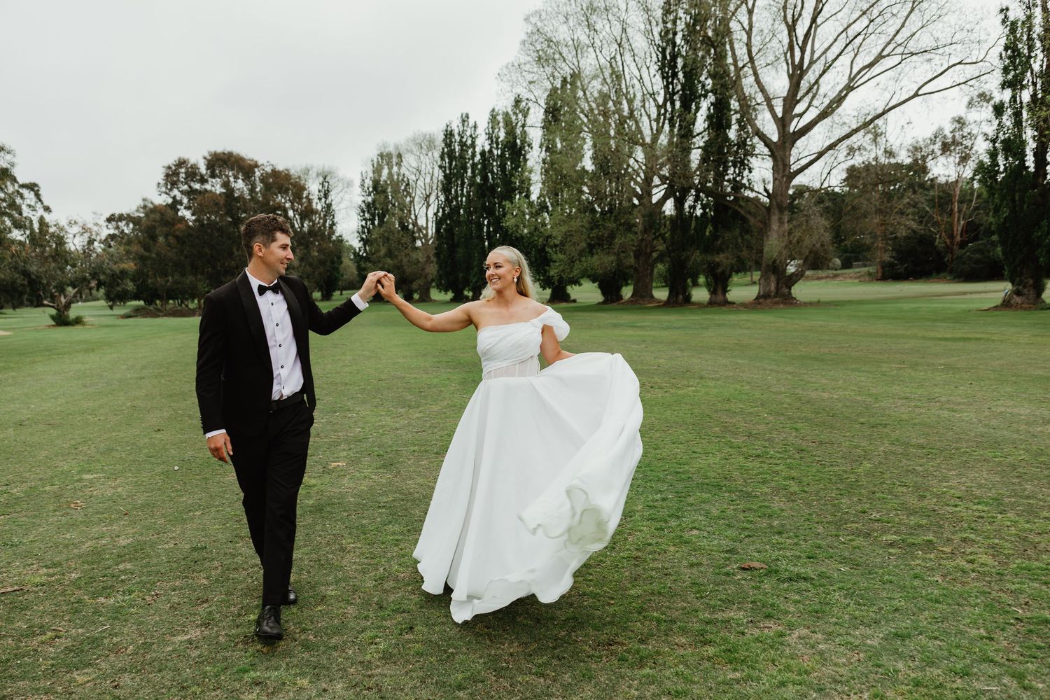 Bride and groom walk hand in hand on green lawn, her white gown flowing in wind.