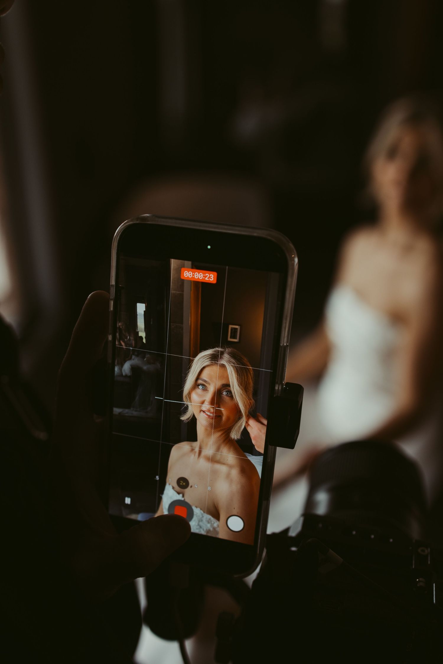 Bride being filmed on smartphone during wedding preparations, blonde woman in white dress captured on phone screen.